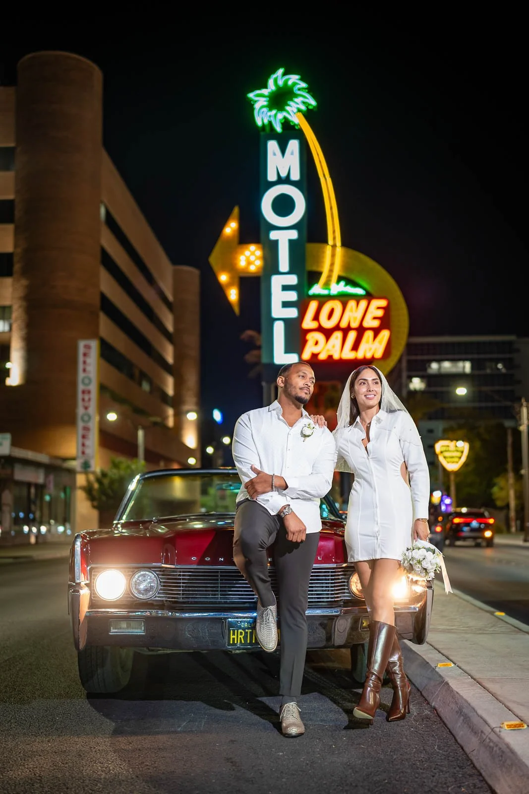 A couple dressed in white, standing next to a vintage red car on a city street at night. The woman is holding a bouquet of flowers, and both are looking away from the camera. Neon signs in the background read "LONE PALM" and "MOTEL" with a palm tree 