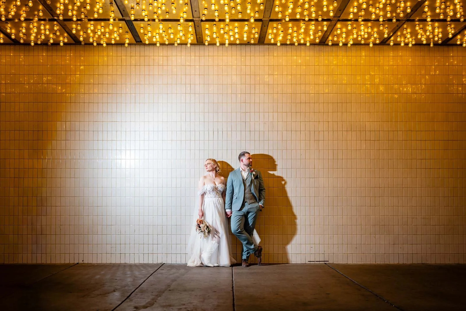 A couple in wedding attire standing against a light-colored tiled wall illuminated by gold string lights from the ceiling, with shadows cast on the wall.