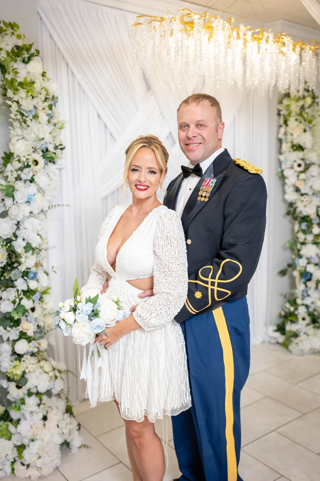 A bride and groom posing together at their wedding, with the bride holding a bouquet of white and light blue flowers, standing in front of a floral arch and white draped backdrop with a crystal chandelier hanging above.