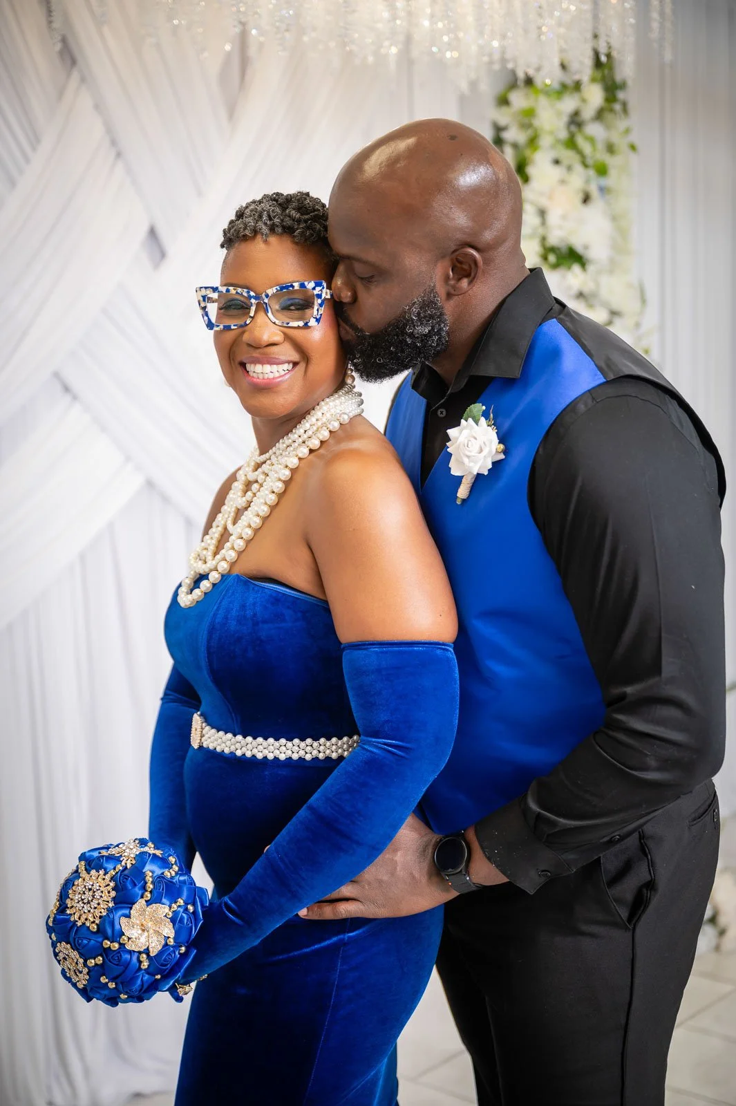 A couple dressed in matching royal blue outfits, with the woman wearing pearl jewelry and glasses, and holding a decorative blue purse, sharing an intimate moment at their wedding.