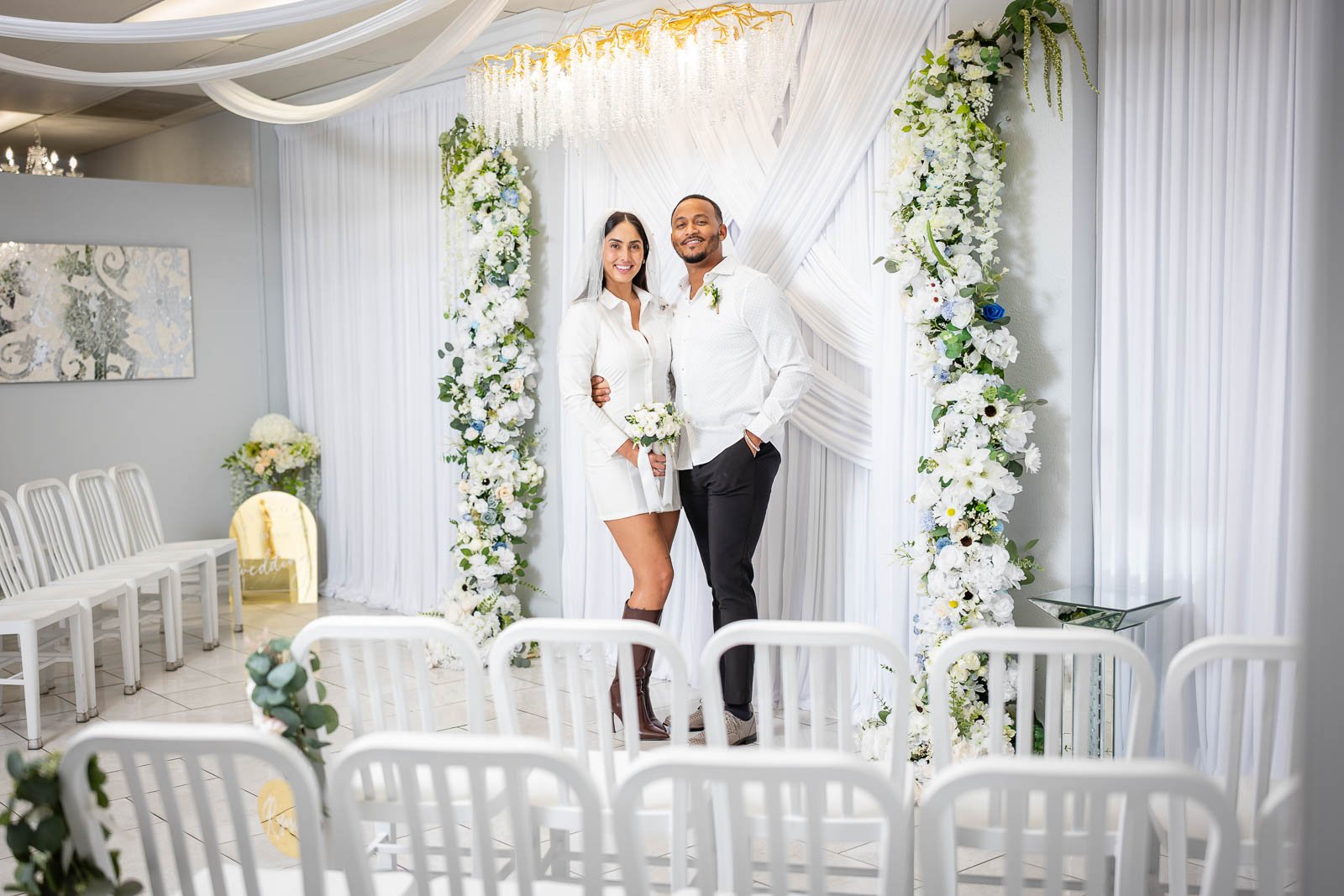 A bride and groom standing together at their wedding ceremony, smiling in front of a decorated backdrop with white flowers and drapery.