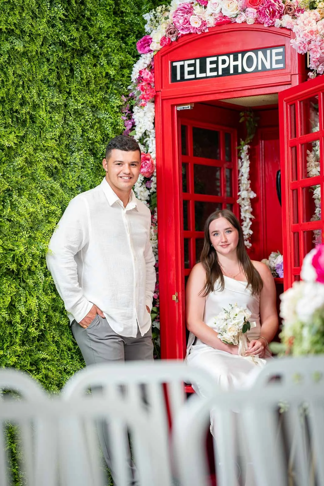 A young man and woman at a wedding, with the woman seated inside a red telephone booth decorated with pink and white flowers and the man standing outside. The woman holds a bouquet of flowers, and the background features a green wall of foliage.