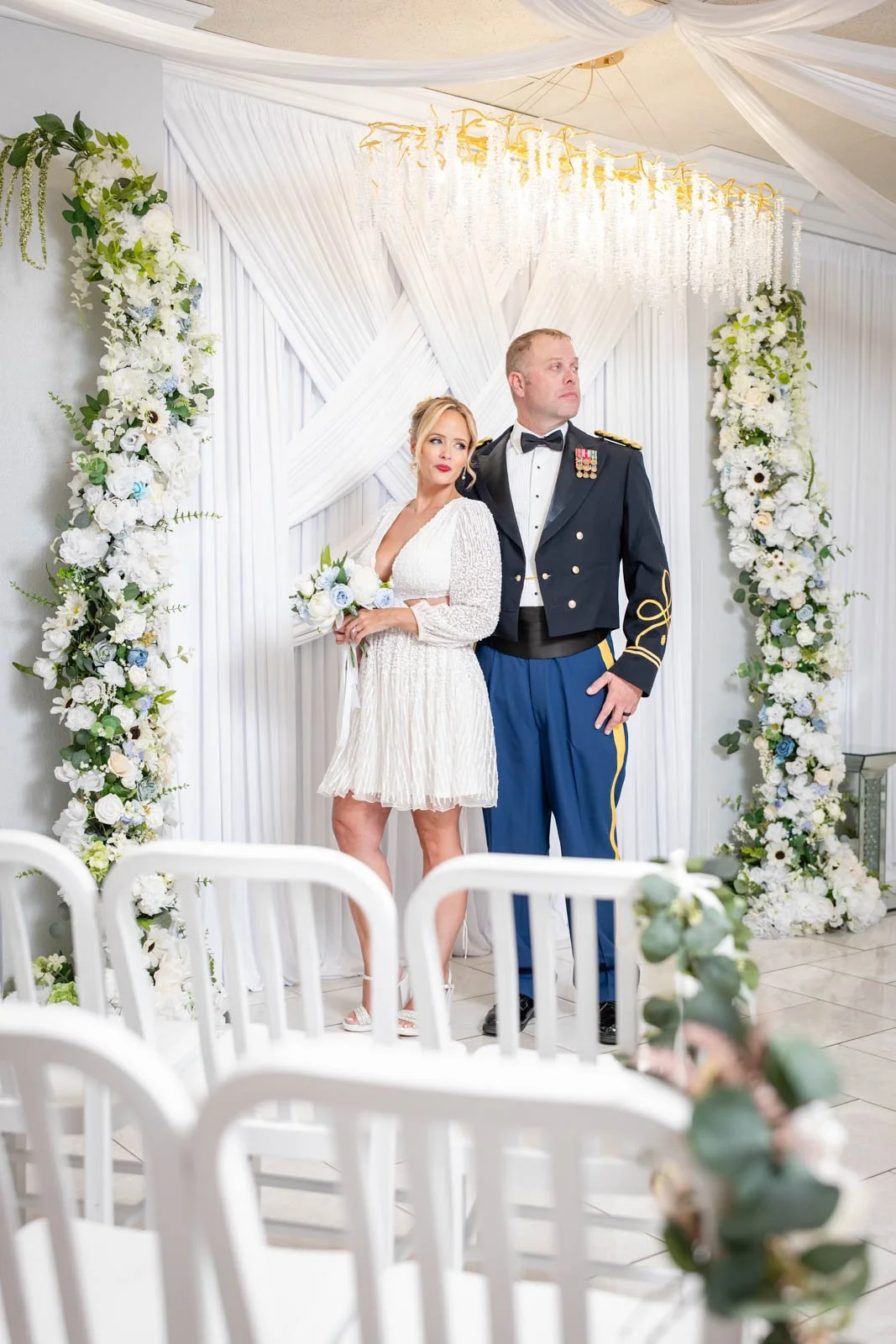 A bride and groom standing under a floral arch during a wedding ceremony. The bride is in a white dress holding a bouquet, and the groom is in a military uniform. The background is decorated with white drapery and floral arrangements.