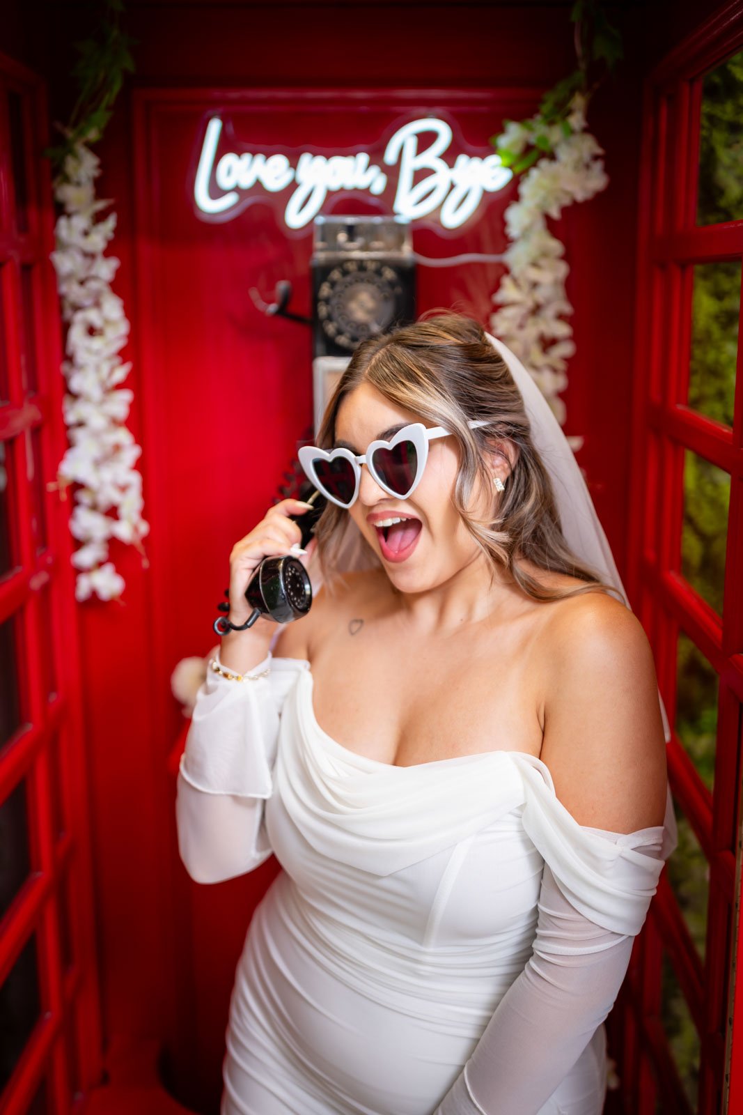 Woman in a white off-the-shoulder wedding dress wearing heart-shaped sunglasses, holding a vintage-style telephone receiver, inside a red telephone booth decorated with white flowers and a neon sign that says 'Love You, Bae'.