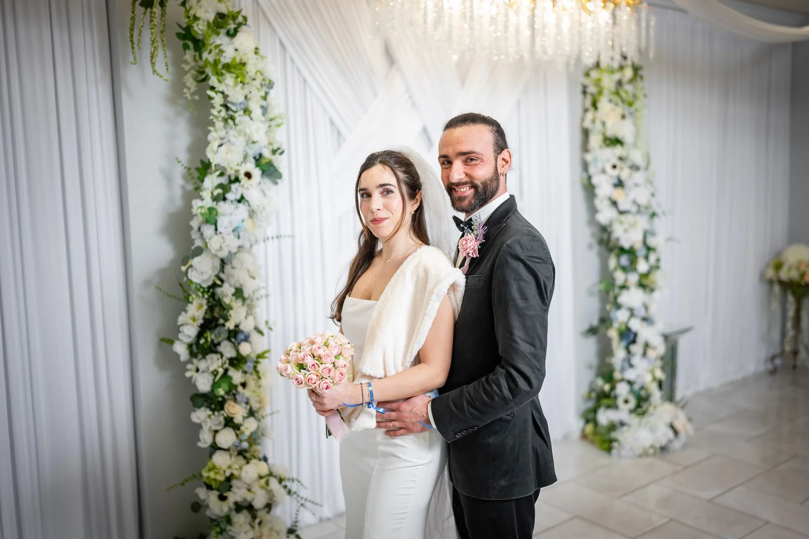 A bride and groom standing together during their wedding ceremony at the Say I Do Chapel with a floral arch and white drapery in the background.