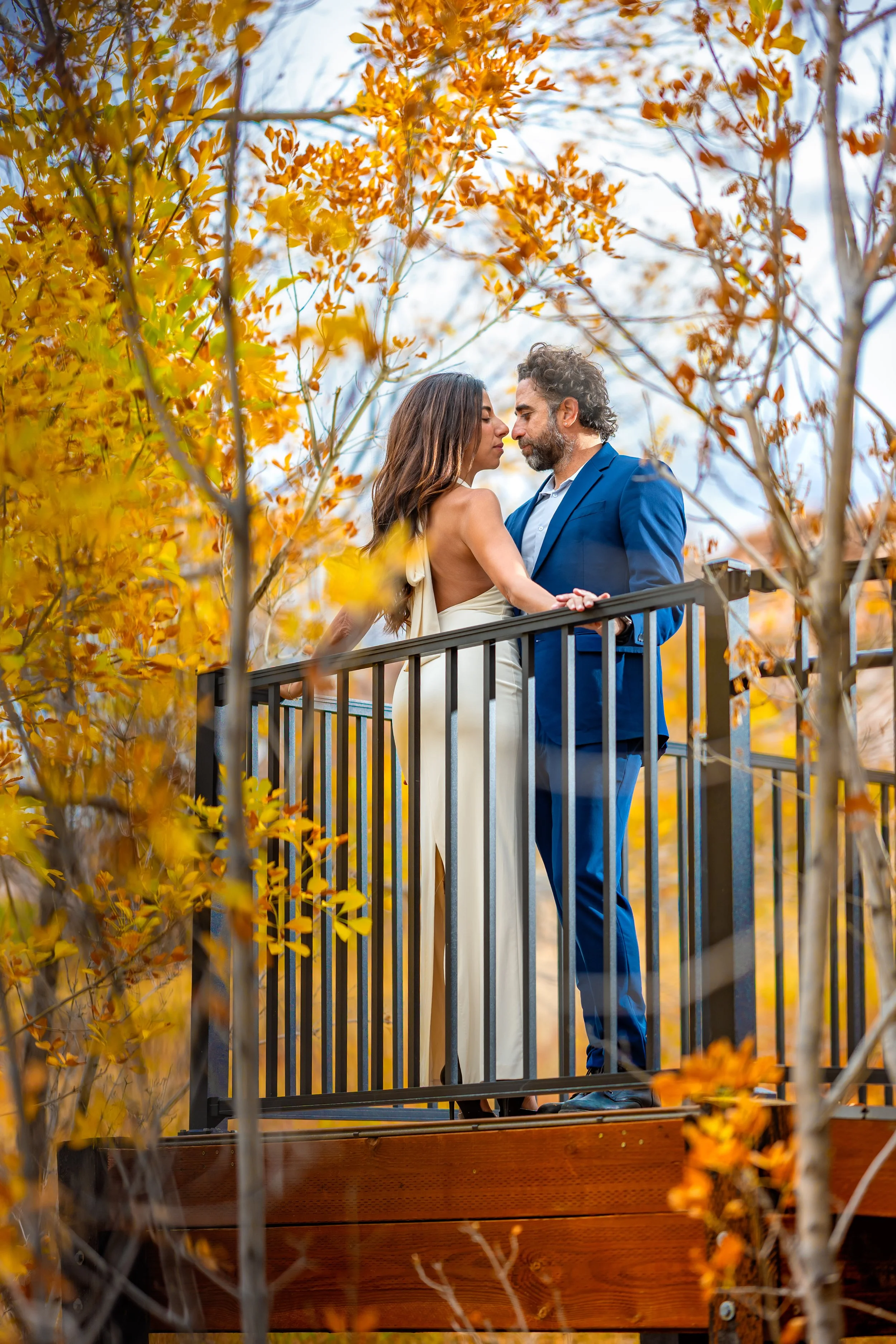 A couple standing on a balcony surrounded by golden autumn leaves, leaning close to each other with their foreheads touching, sharing an intimate moment.