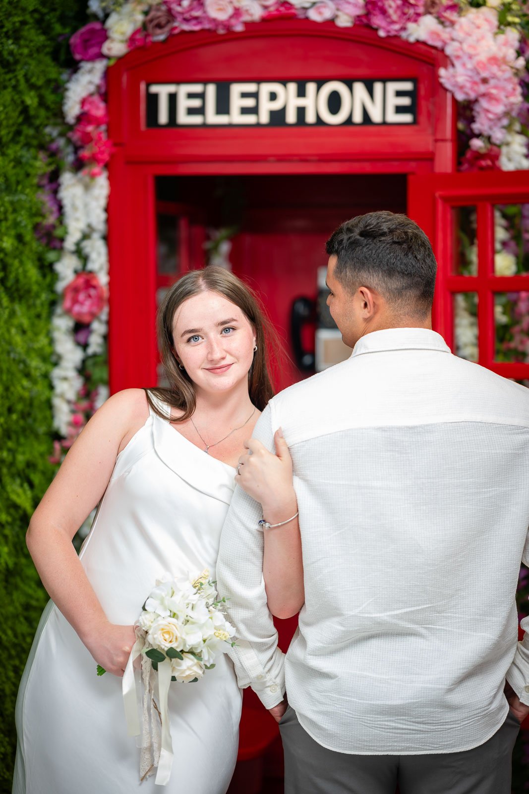 A young woman in a white dress holding a bouquet of white flowers stands beside a young man in a white shirt, with her hand on his shoulder, in front of a red telephone booth decorated with flowers.