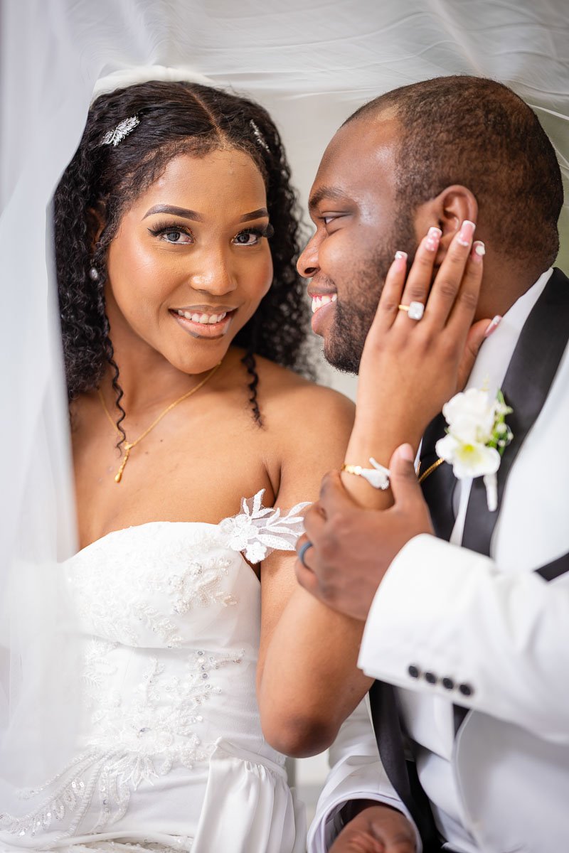 A bride and groom share an intimate moment, with the bride gently holding the groom's face as they smile at each other inside a wedding canopy.
