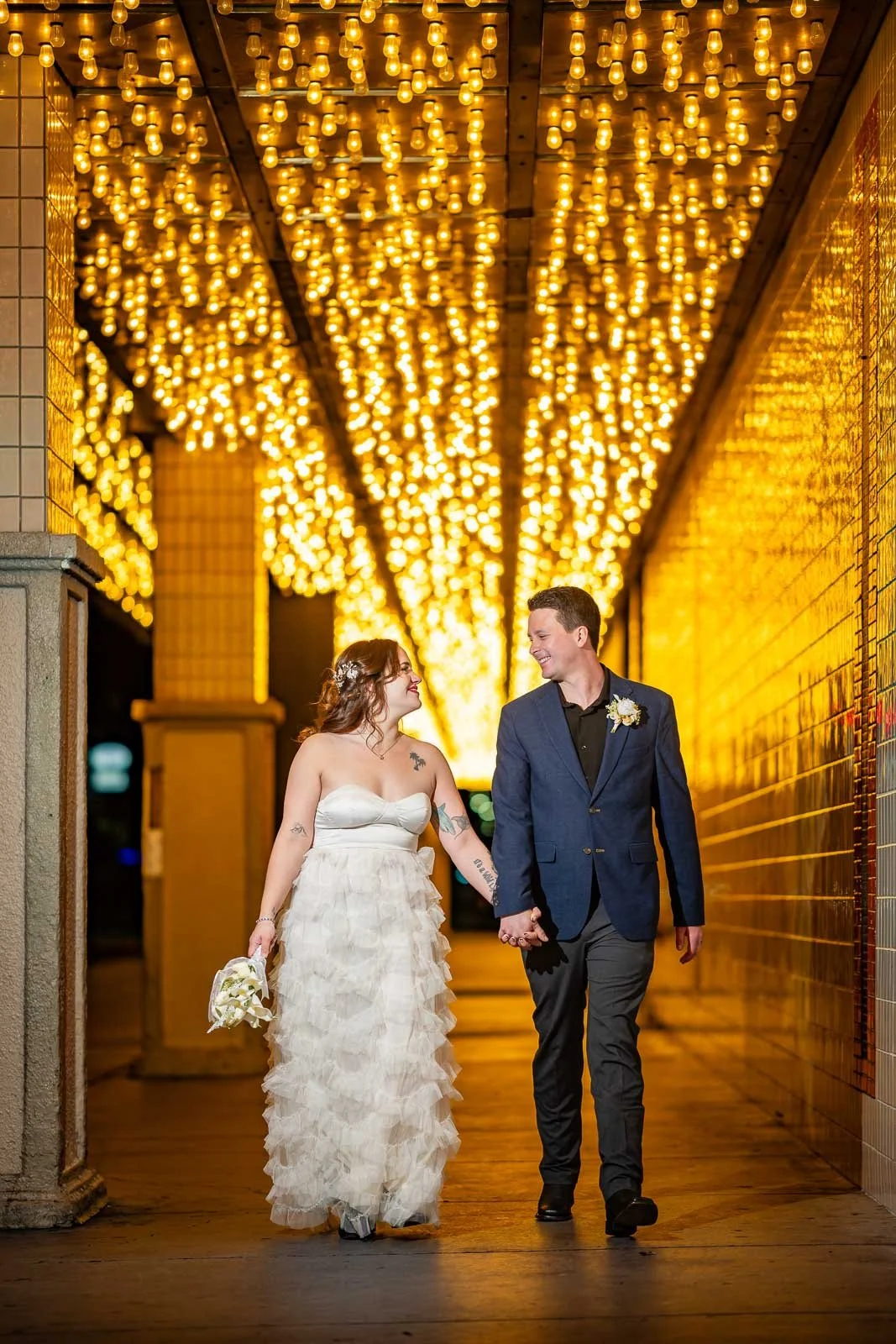 A newlywed couple is walking hand in hand under a tunnel of warm yellow lights at night. The bride is wearing a strapless white wedding dress with ruffles, holding a bouquet, and has tattoos on her arms. The groom is dressed in a navy blazer, black s