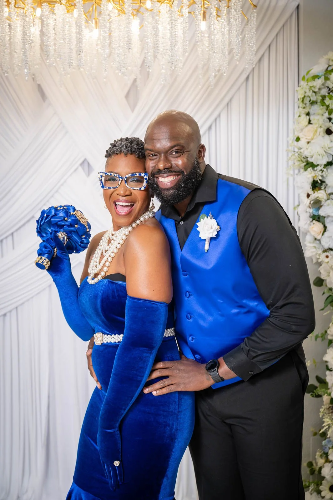 A joyful couple dressed in blue and black posing at a wedding or celebration. The woman is wearing a royal blue velvet dress with matching gloves and accessories, and the man is in a blue vest with black shirt and pants. They are smiling happily with