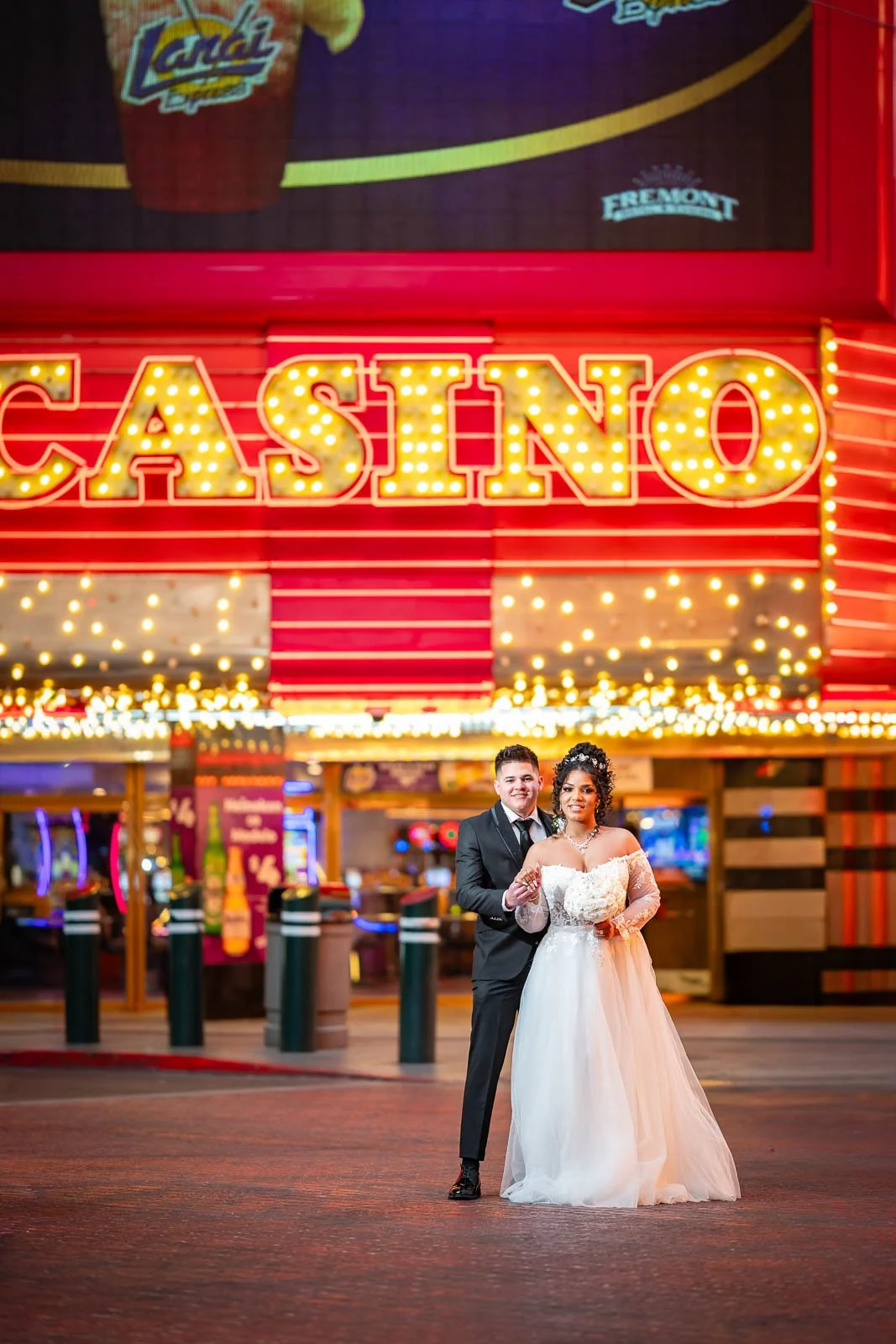 A wedding couple stands outside a brightly lit casino entrance at night. The groom is dressed in a black tuxedo with a black tie, and the bride is in a white wedding gown with lace details. Behind them, a large illuminated sign reads 'CASINO' with ye