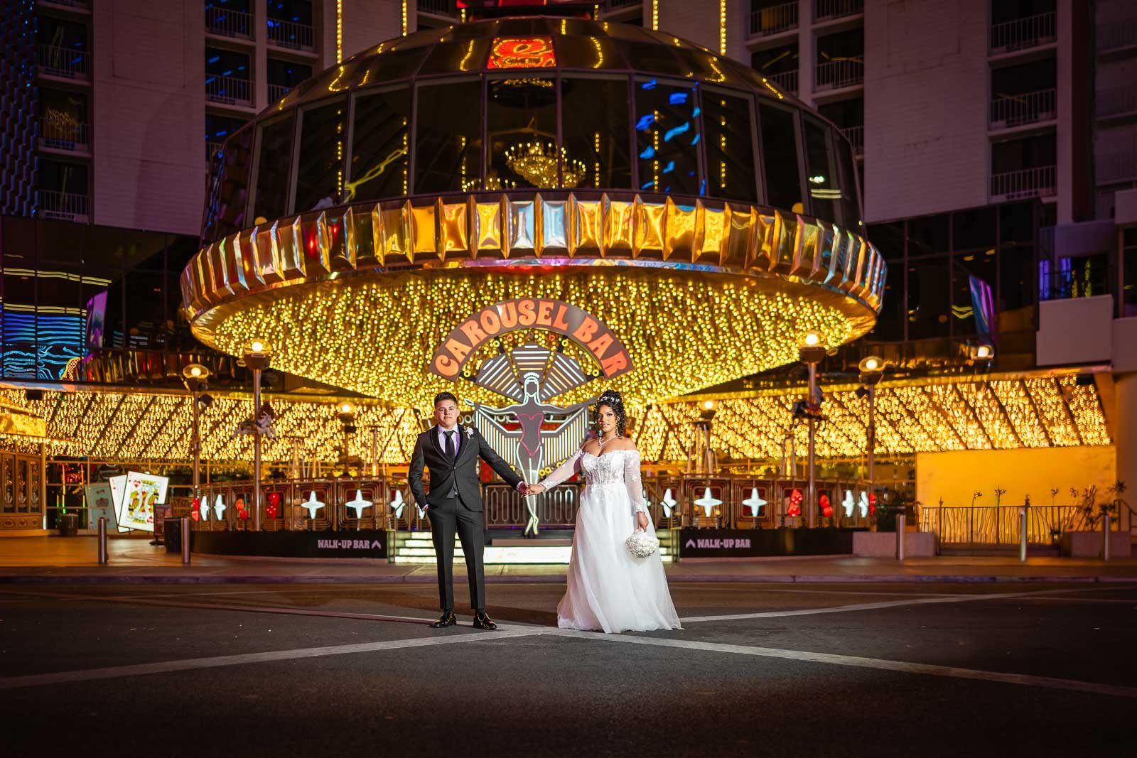 A bride and groom holding hands in front of a brightly lit carousel at night. The bride wears a white wedding gown and holds a small bouquet, while the groom is dressed in a black tuxedo. The carousel behind them has yellow lights and a sign that rea