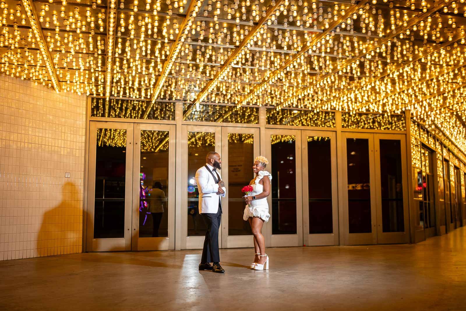 A couple in formal attire standing and talking under a ceiling of yellow string lights outside a building with glass doors at night in downtown Las Vegas.