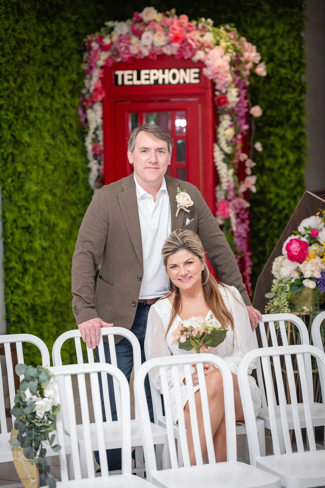 A man and woman posing at a wedding reception with a red British telephone booth decorated with pink and white flowers in the background. The woman is sitting with a bouquet of flowers, and the man is standing behind her.