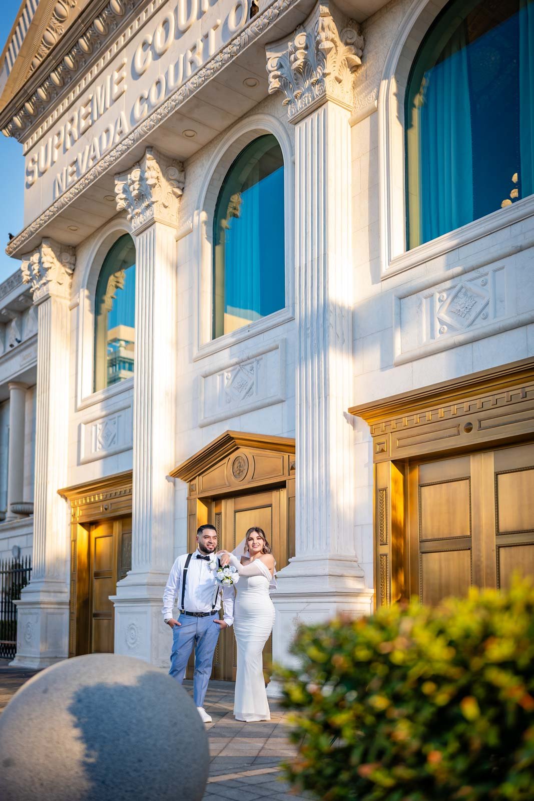 A newlywed couple standing outside an ornate building with large arched windows, white marble facade, and wooden doors. The bride is in a white dress holding a bouquet, and the groom is in a white shirt with suspenders and gray pants. The scene is il