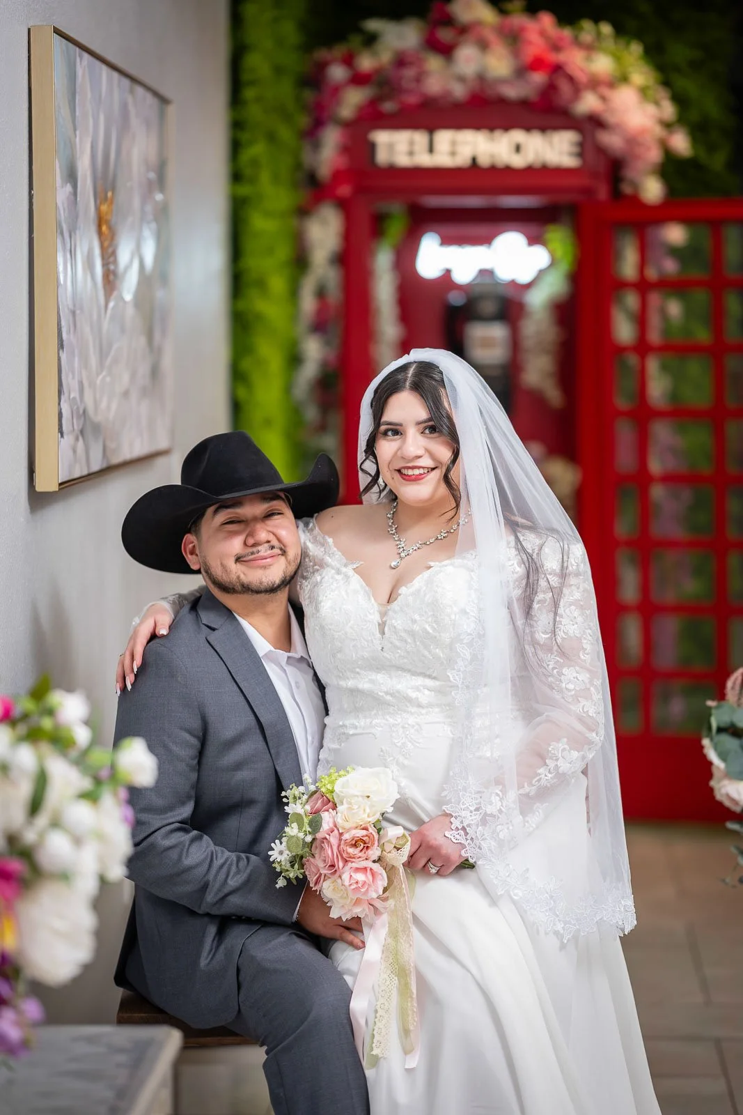 A bride and groom posing indoors. The groom is sitting, wearing a gray suit and a black cowboy hat, holding a bouquet of pink and white flowers. The bride is standing beside him, wearing a white lace wedding dress with a veil and jewelry. They are sm
