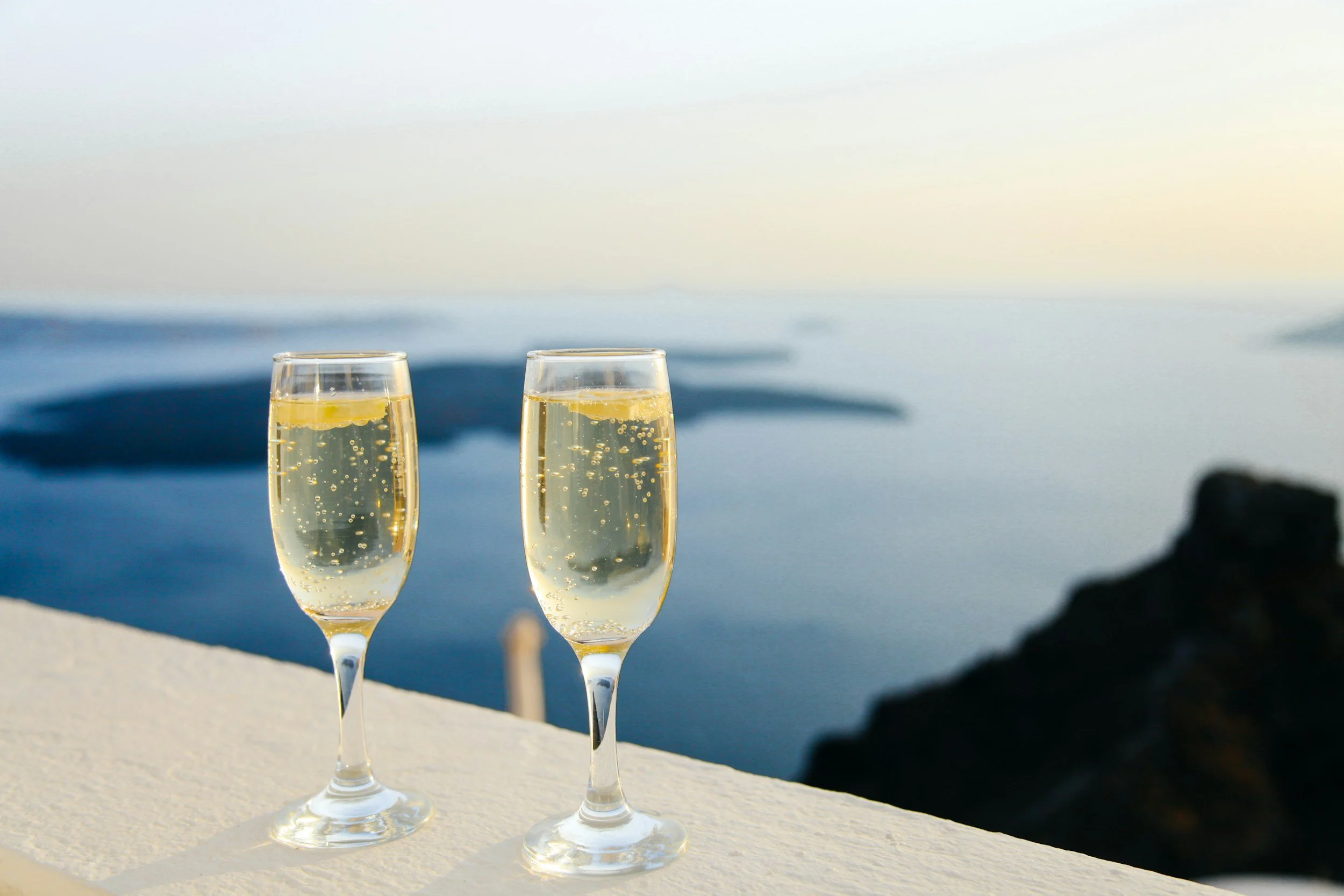 Two glasses of champagne with lemon slices on a ledge near the ocean at sunset.