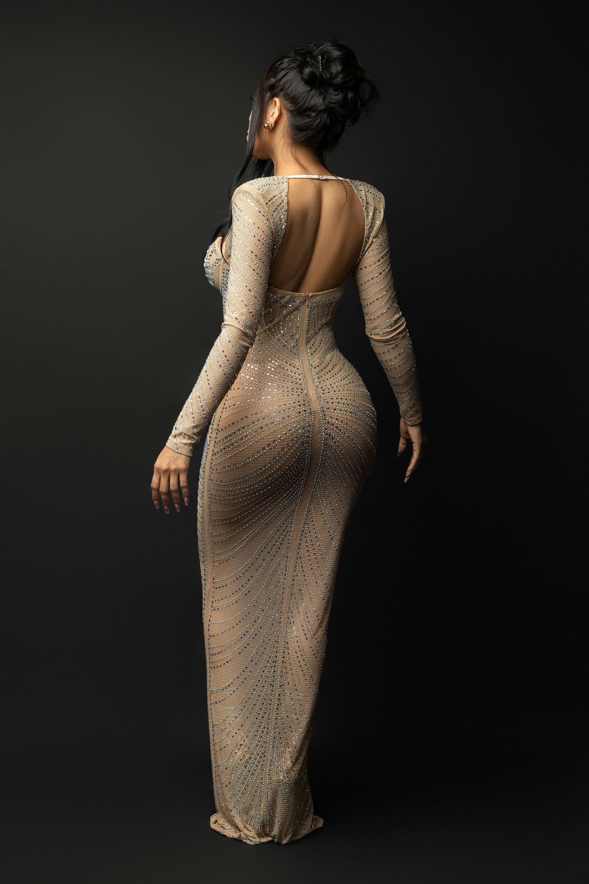 A woman in a fitted, long, shimmering gown with a backless design, standing against a black background, with dark hair styled in an elegant updo.