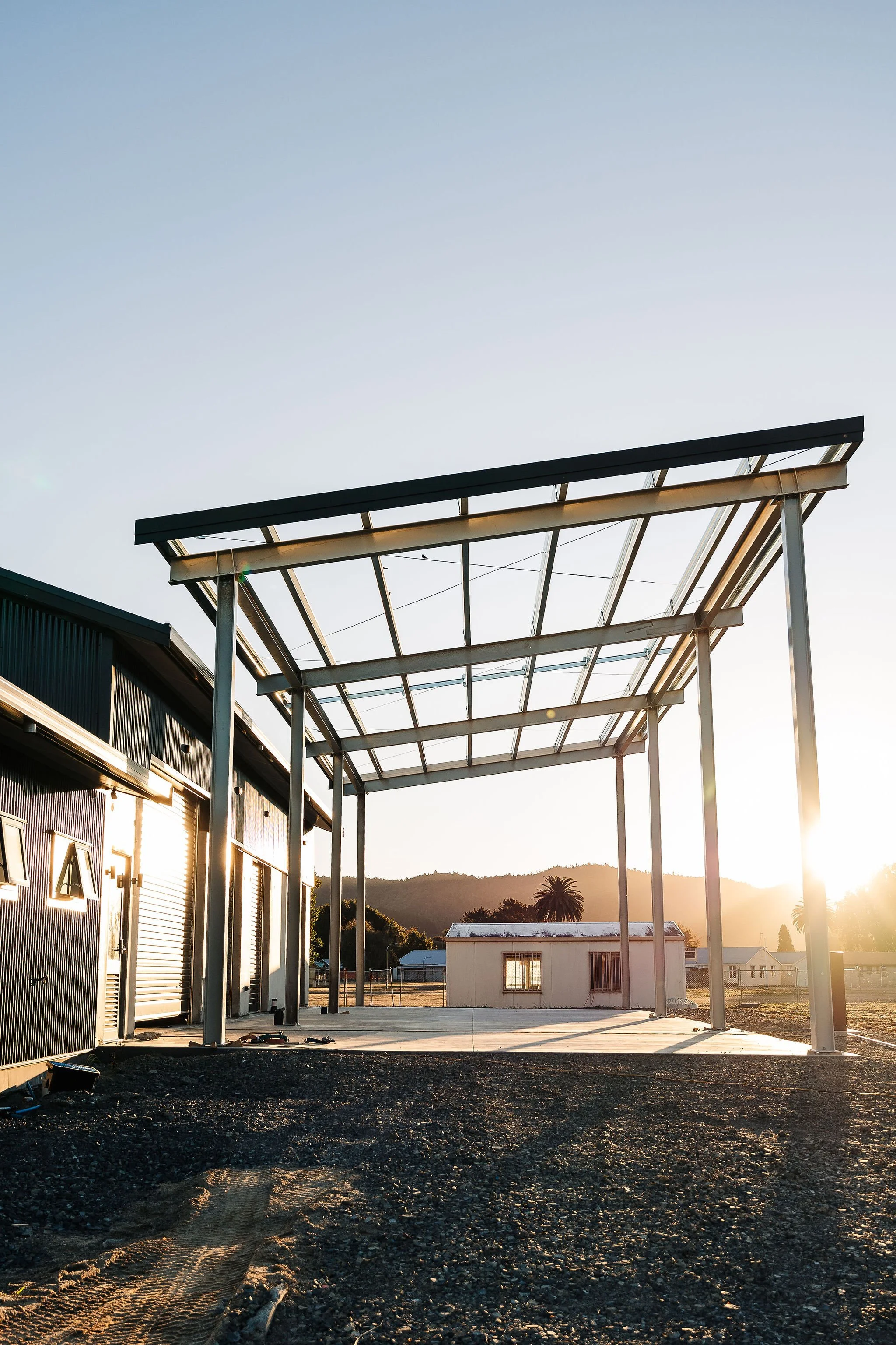 Construction site with a metal structure framework and a partially completed roof, during sunset with distant mountains and palm trees in the background.
