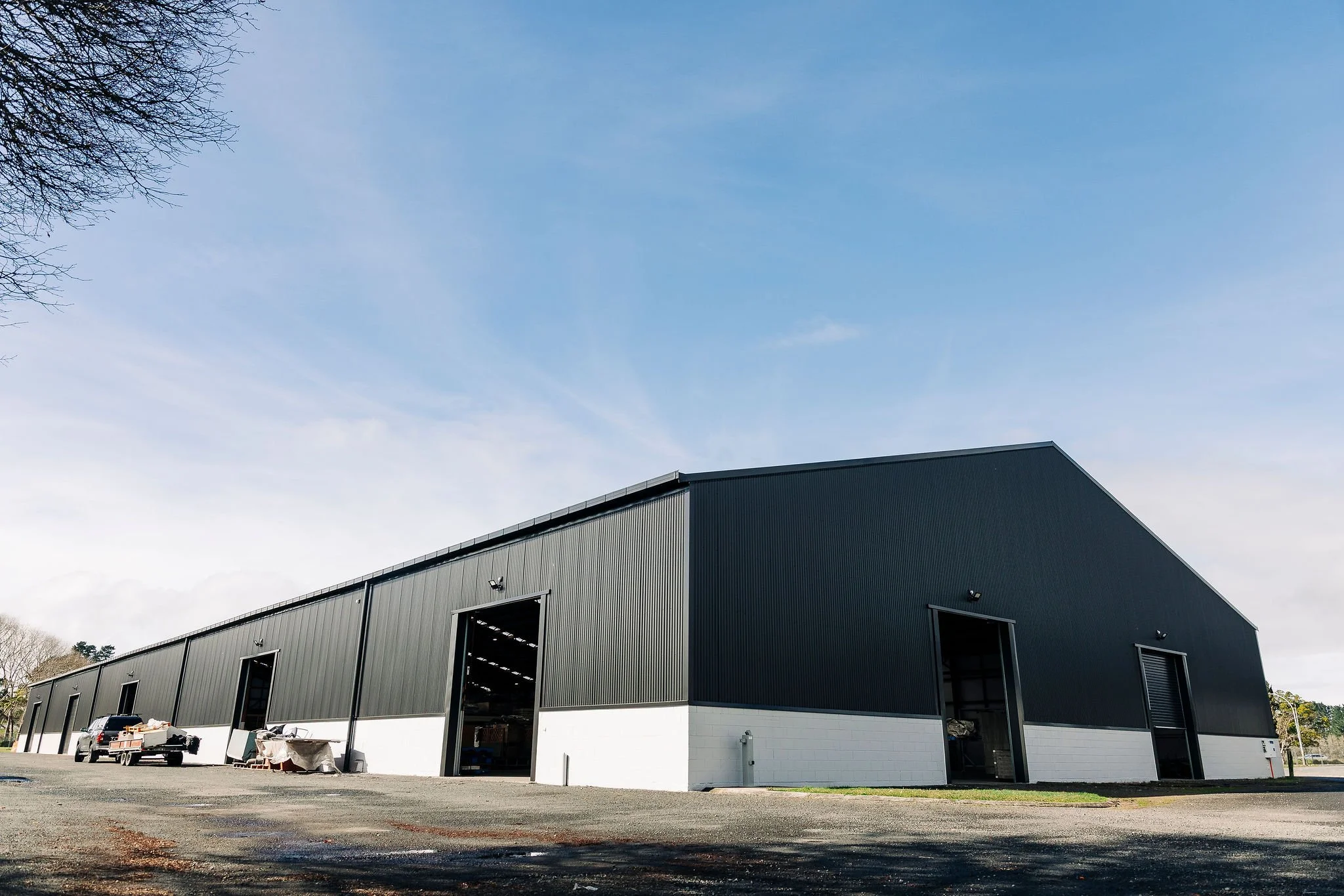 Large gray industrial warehouse with open garage doors, parked vehicles, and construction materials outside, under a clear blue sky.