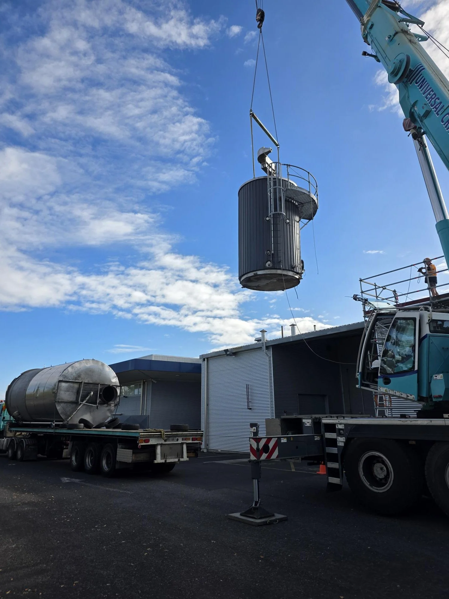 A large crane lifting a cylindrical metal tank at an industrial site, with a person standing on a scaffold nearby and industrial buildings in the background.