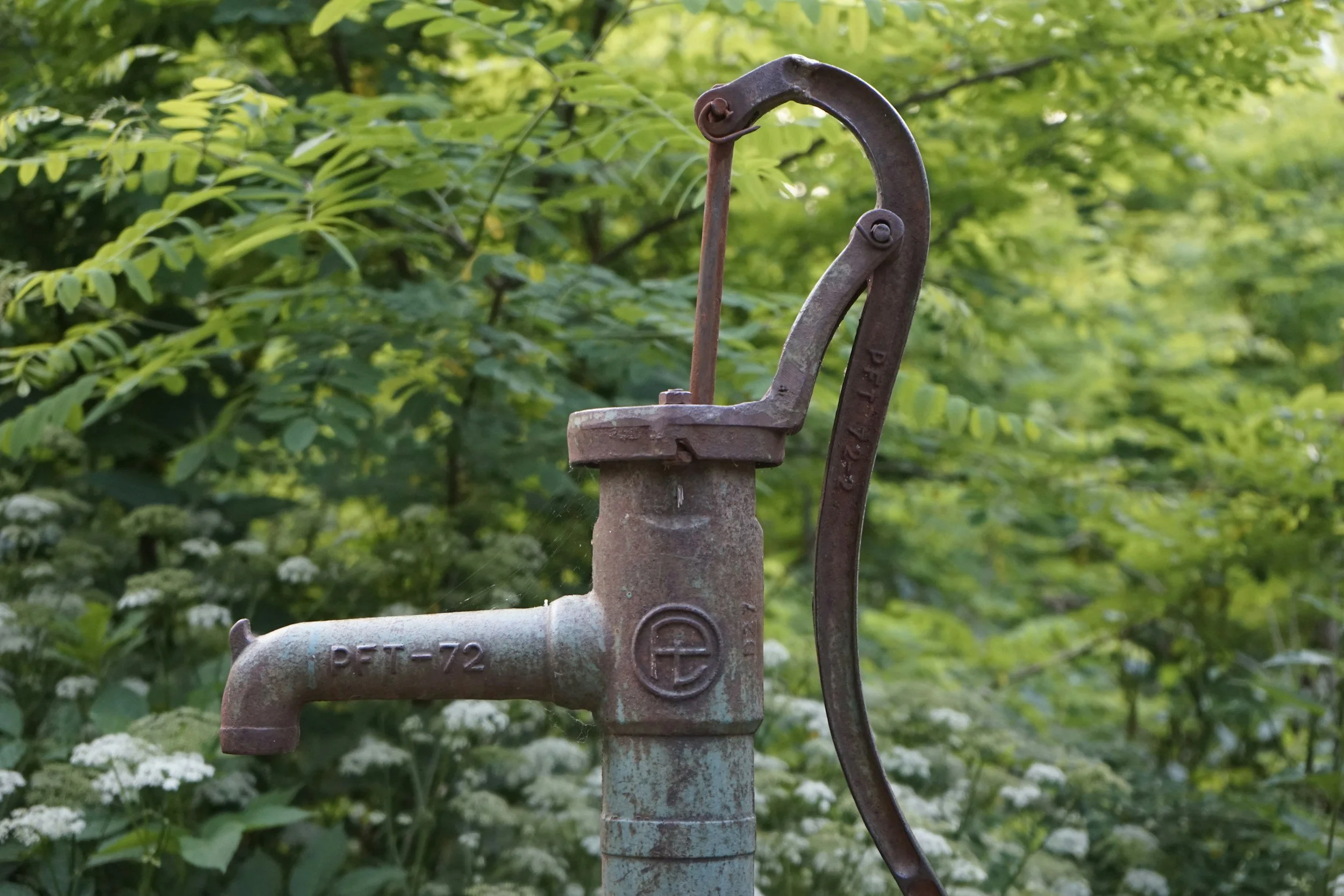 An old rusted hand water pump with green foliage in the background.