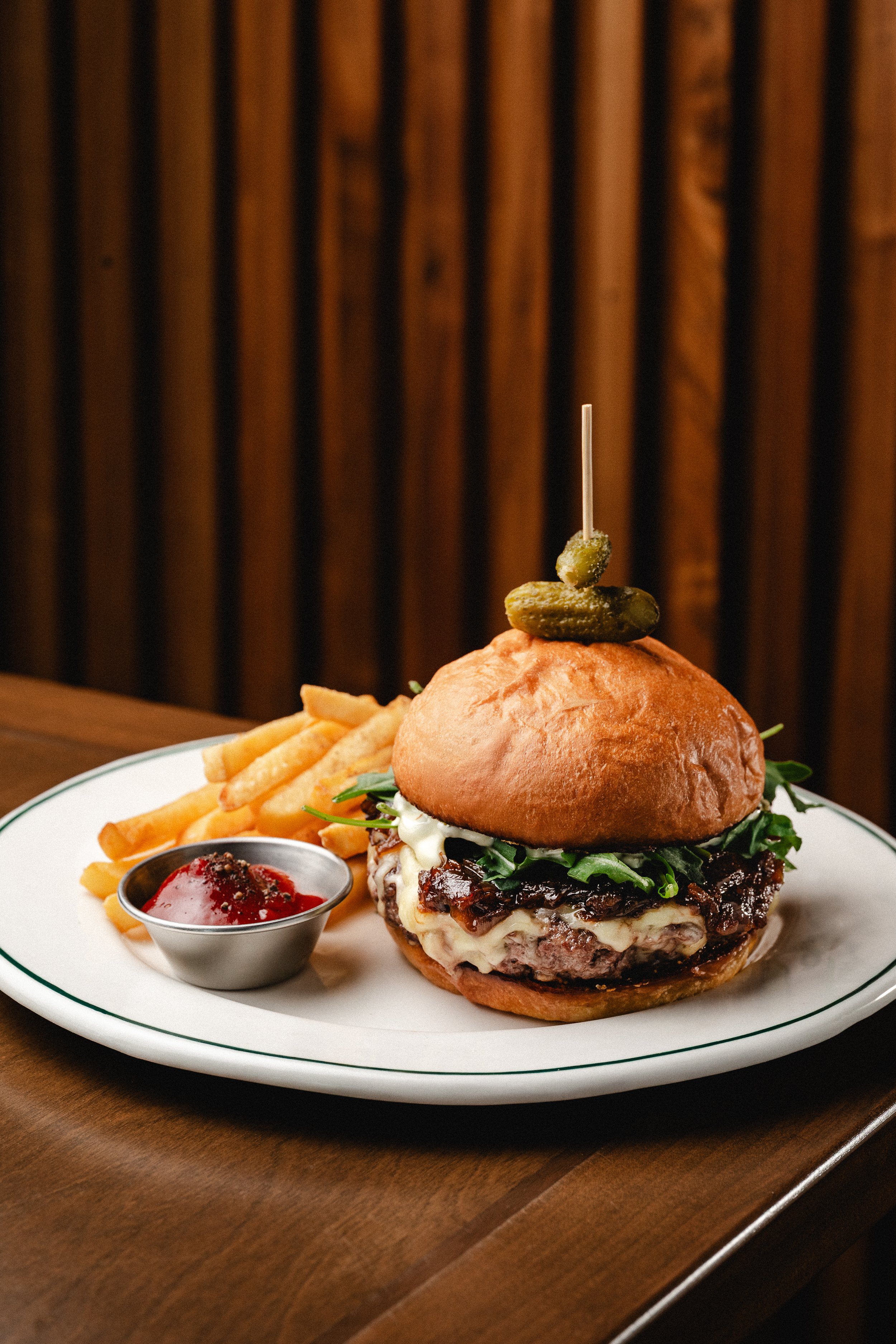 Hamburguesa con papas fritas, salsa y pepinillos en un plato blanco sobre una mesa de madera.