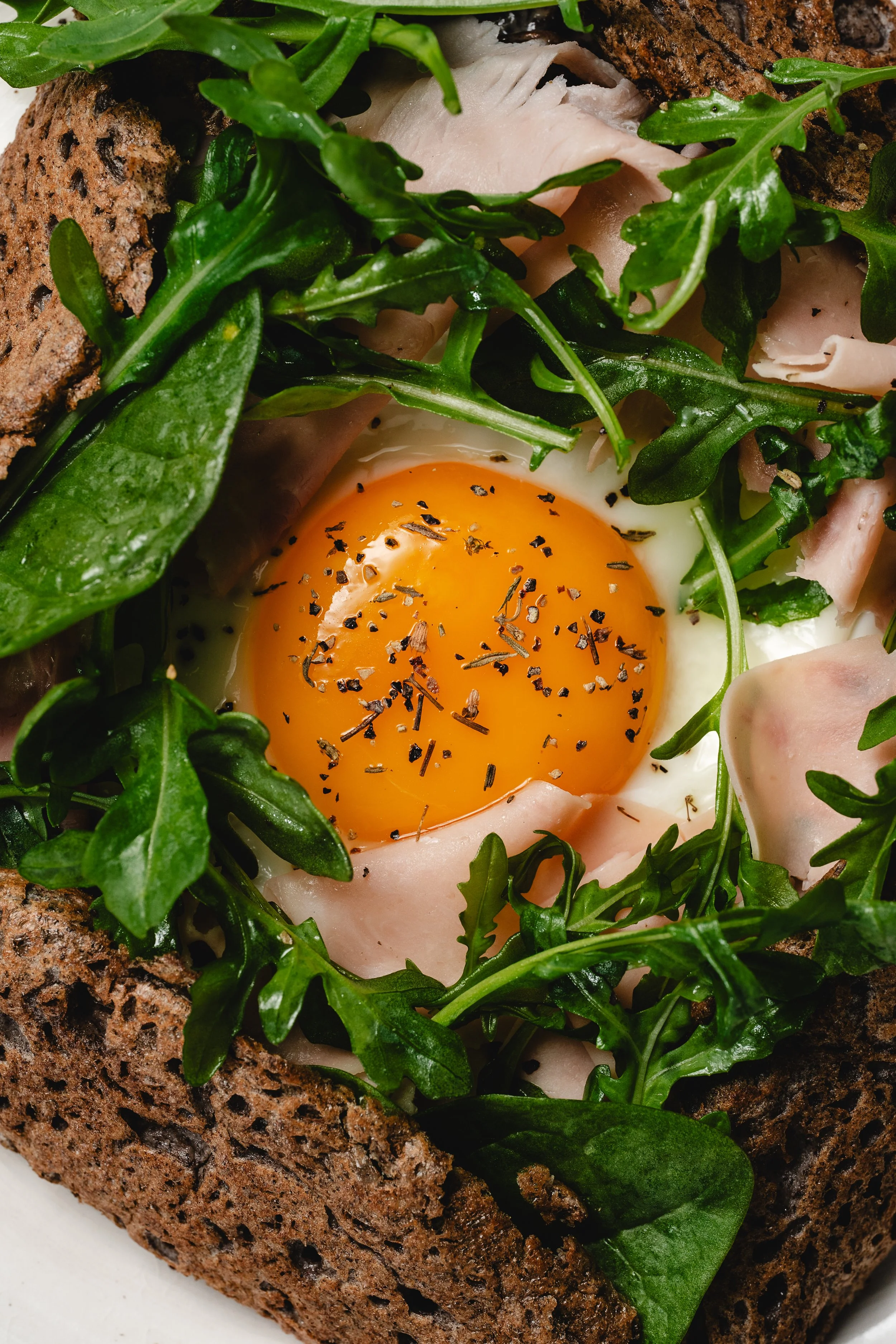Huevo cocido en un pan de centeno con espinacas, arúgula, rebanadas de jamón y pimienta negra molida