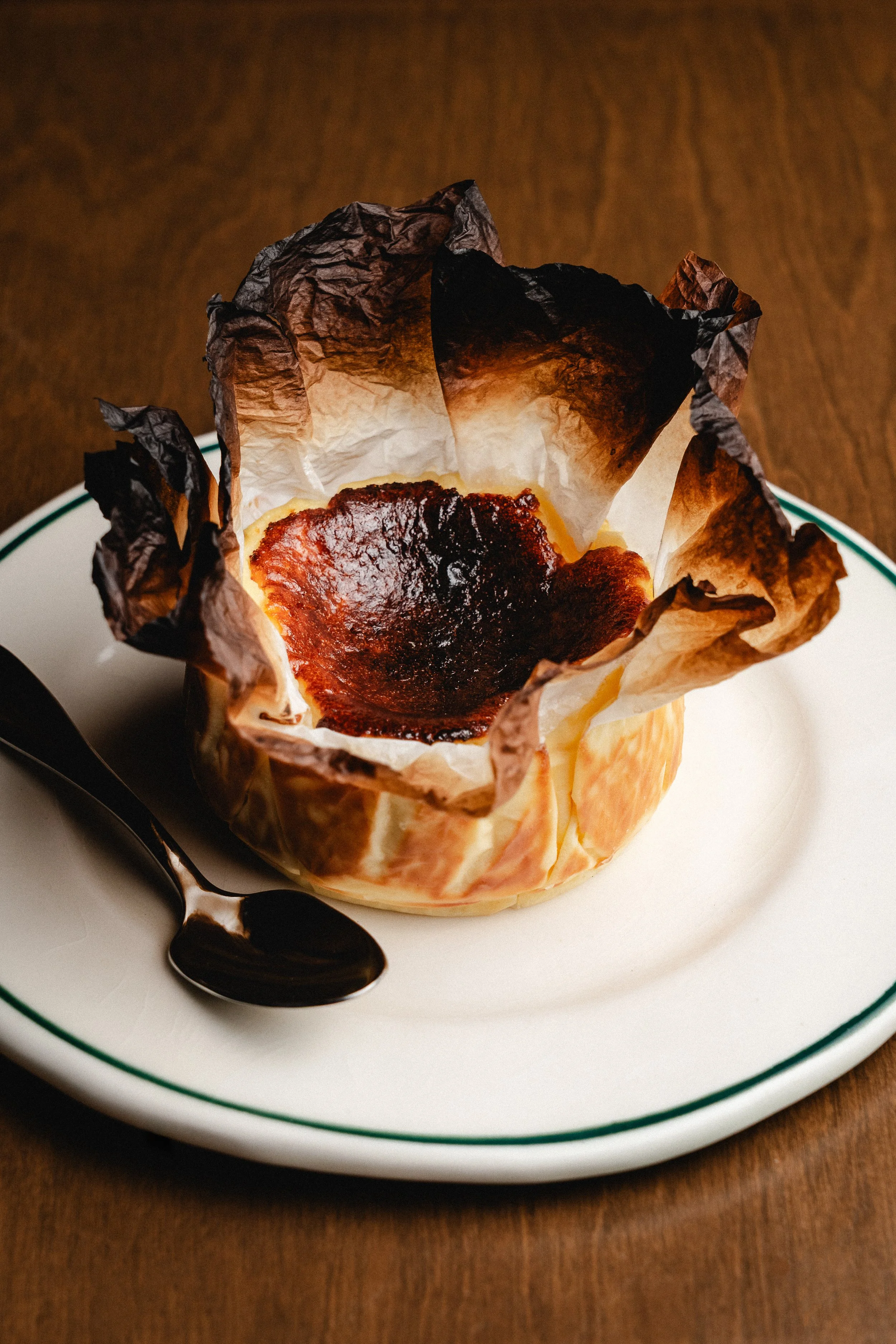 Una copa de vino con la parte superior quemada y parcialmente cubierto con papel de seda quemado, sobre un plato blanco con borde verde y un cuchara negra al lado, sobre una mesa de madera.