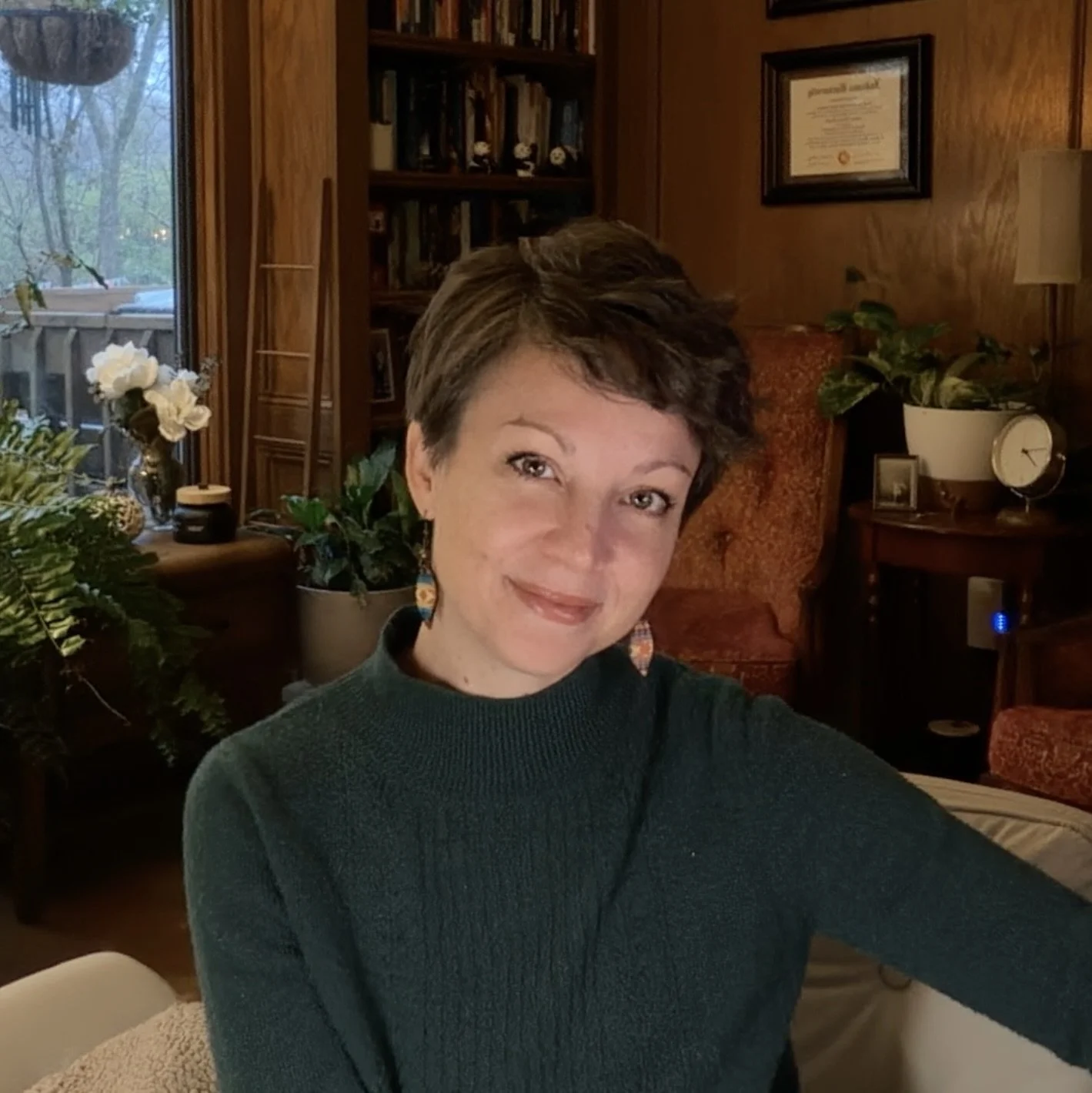 A woman with short brown hair wearing a dark green sweater is sitting in a cozy living room with wooden walls and a collection of plants, books, and framed photos and certificates in the background.