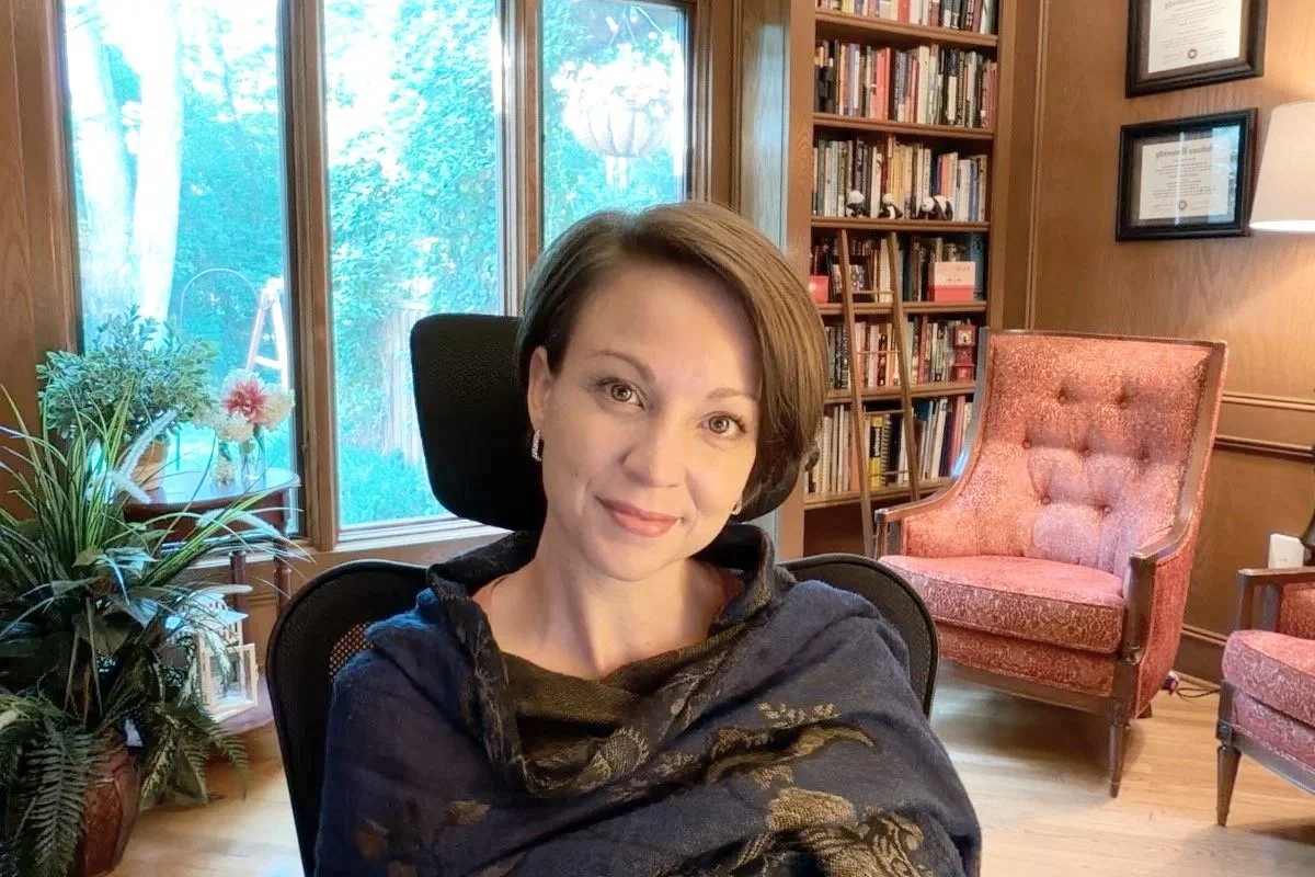 A woman with short brown hair and earrings sitting in an office chair in a cozy room. Behind her are bookshelves filled with books, a window with greenery outside, and a red armchair. There are framed diplomas on the wall and a lamp.