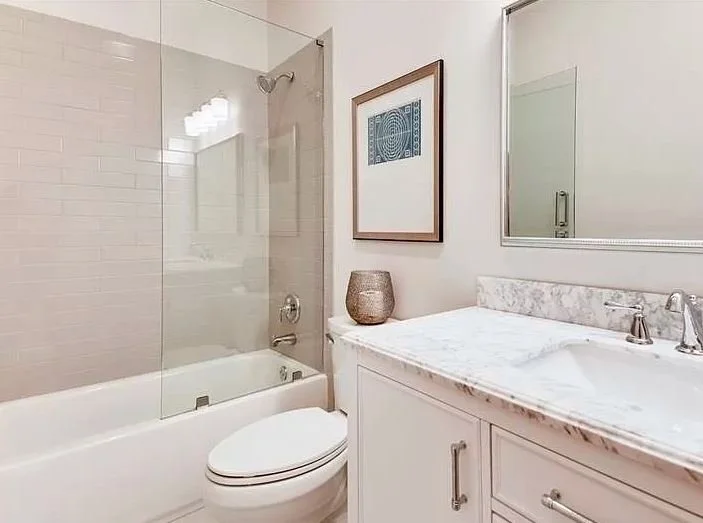 Bathroom featuring a bathtub with a glass shower door, a toilet, a marble countertop with a sink, a framed artwork, and a decorative vase on the countertop.