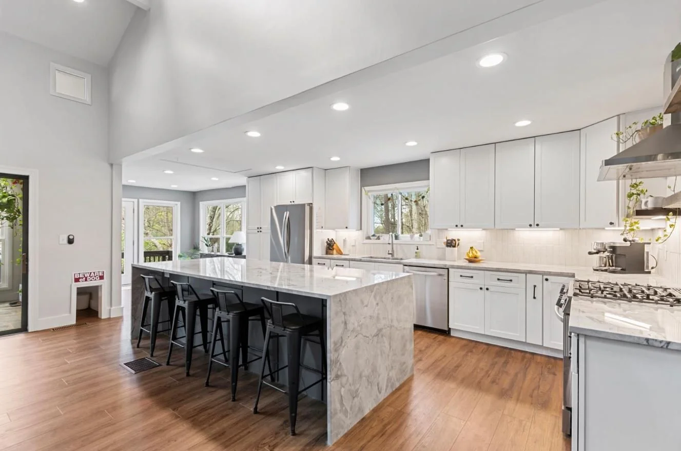 Modern kitchen with white cabinets, marble island, stainless steel appliances, and wooden flooring, with windows showing trees outside.