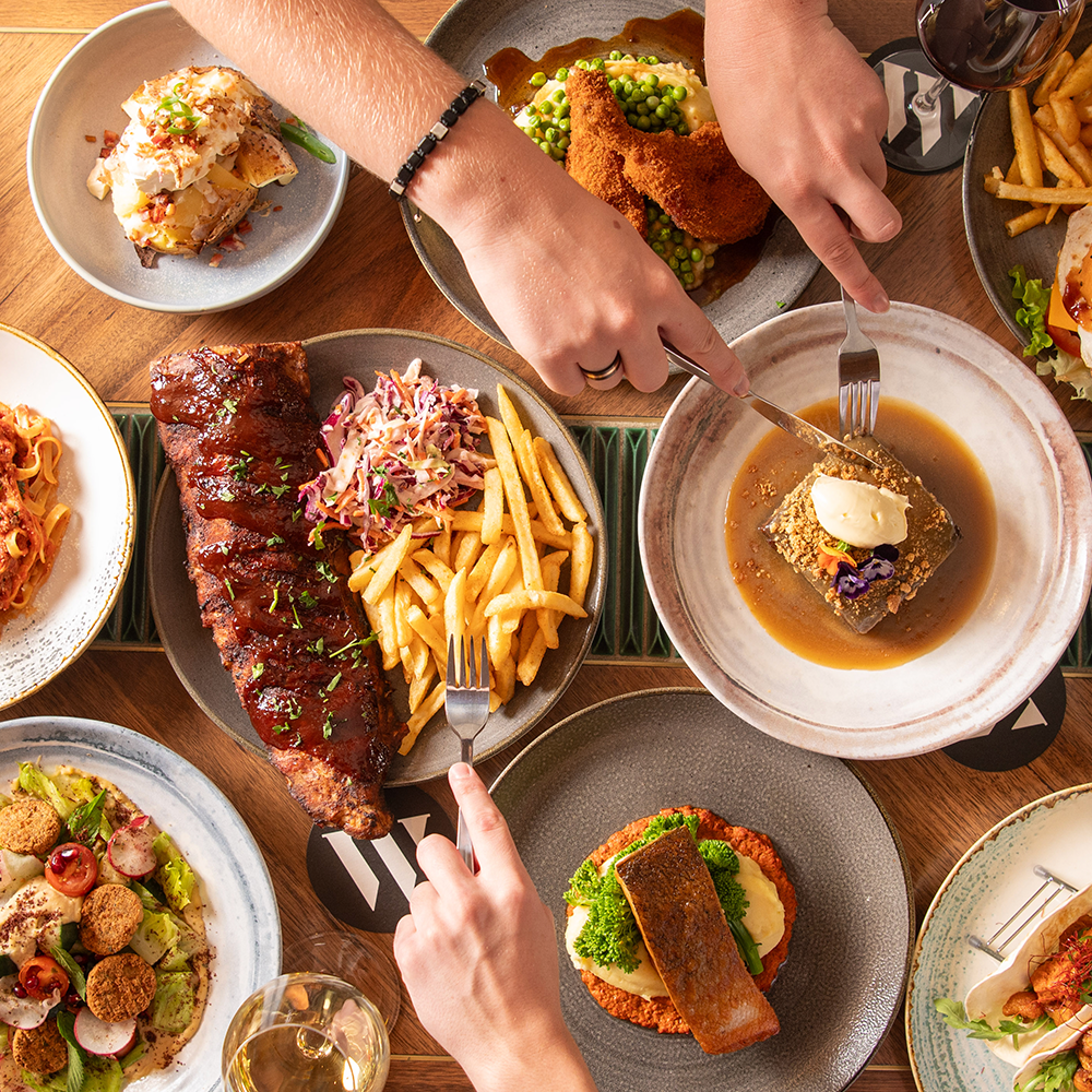 A variety of plated dishes on a wooden table, including French fries with ribs and coleslaw, a meatloaf with gravy, a potato casserole, a salad with croutons, and a drink of red wine.