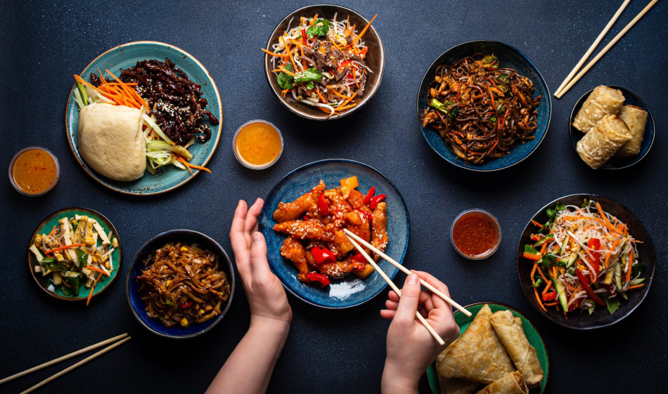 An assortment of Asian dishes on a dark table, including spring rolls, noodles, stir-fried vegetables, and rice, with small bowls of sauces, and chopsticks.