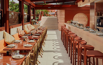 Upscale restaurant with a long bar on the right and a row of tables with chairs and cushions on the left, large windows, indoor plants, and stairway at the back.