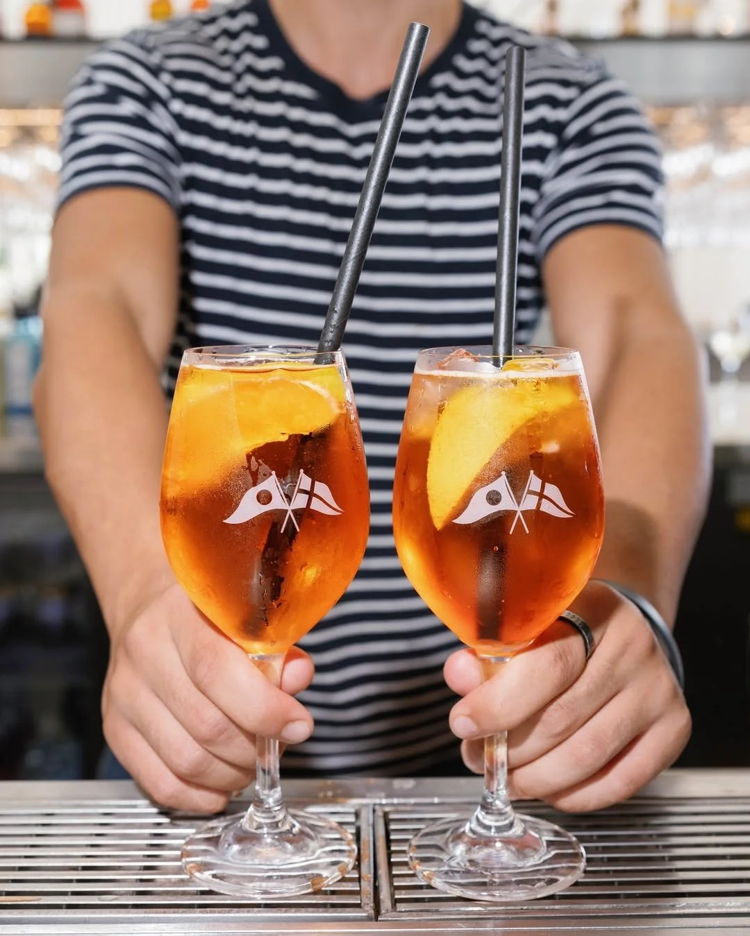 Two glasses of Aperol spritz with ice and lemon slices, held by a person in a striped shirt, bar setting in background.