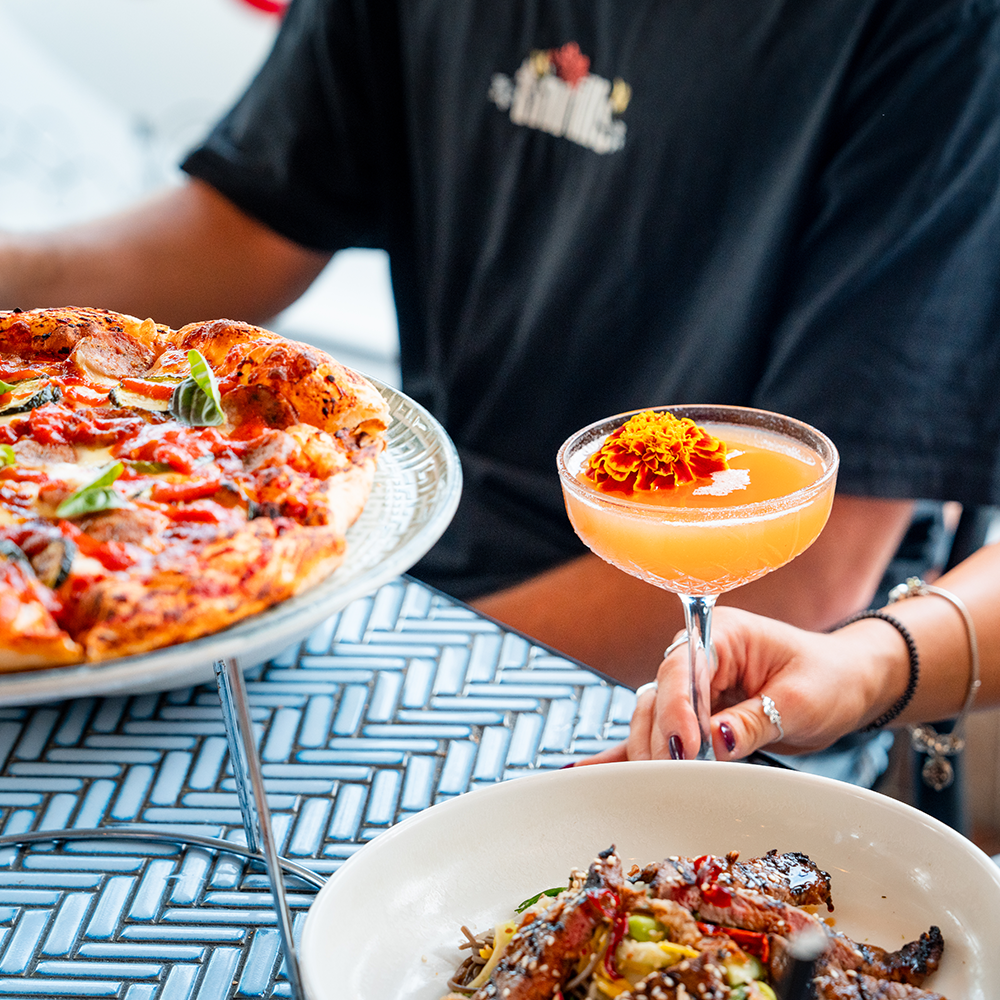 Person holding a pink cocktail with an orange flower garnish, with a pizza on a stand and a plate of food on the table.