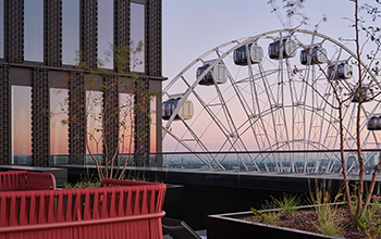 View of a Ferris wheel and a city skyline at sunset from a rooftop balcony with a red metal railing and some plants.
