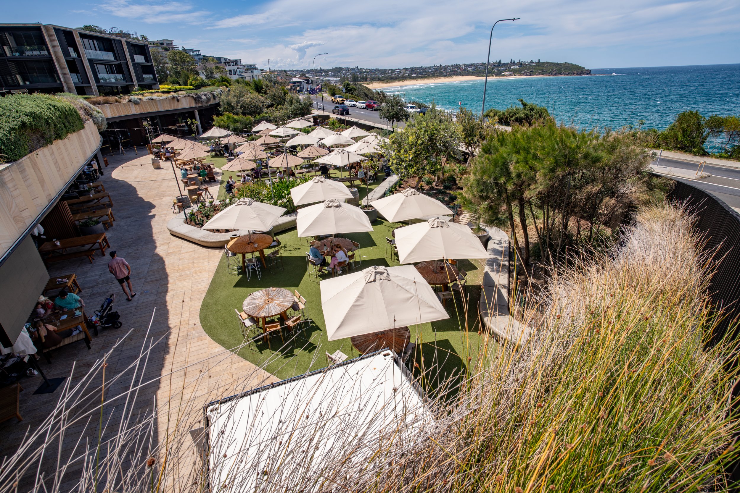 An outdoor dining area on a terrace overlooking the ocean with white umbrellas and tables, modern buildings, a road with cars, and a sandy beach in the background under a partly cloudy sky.