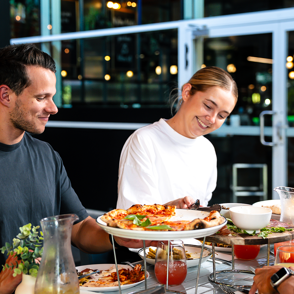 Two people smiling at a table with pizza, drinks, and food in a restaurant.
