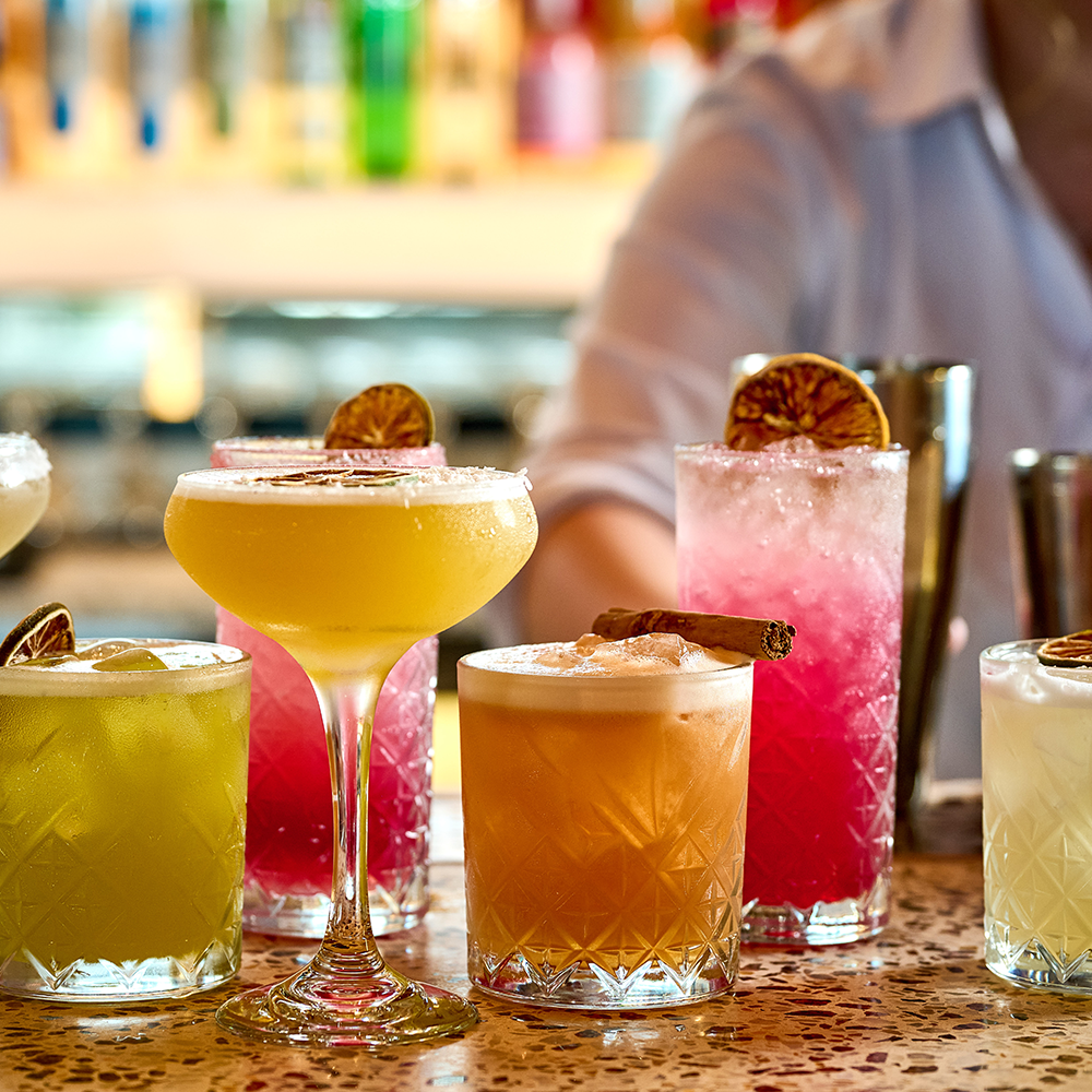 An assortment of colorful cocktails with garnishes like dried citrus slices and cinnamon sticks on a bar counter.