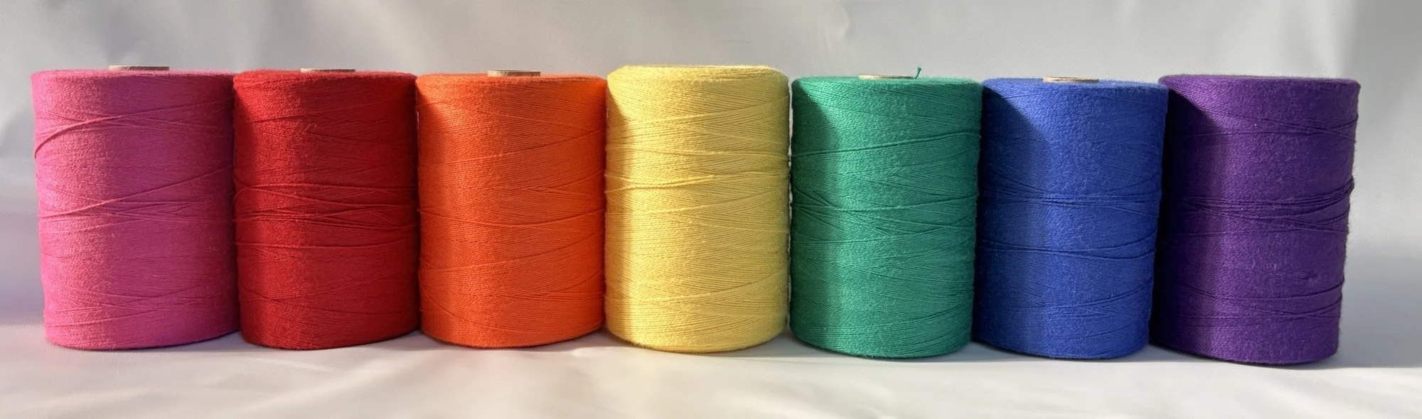 Seven cones of Maurice Brassard cotton weaving yarn in a row, in magenta, red, orange, yellow, green, blue and purple