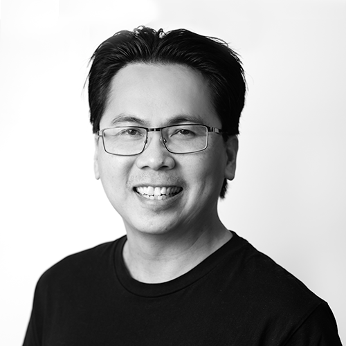 Black and white portrait of a smiling man wearing glasses and a black shirt, facing forward.