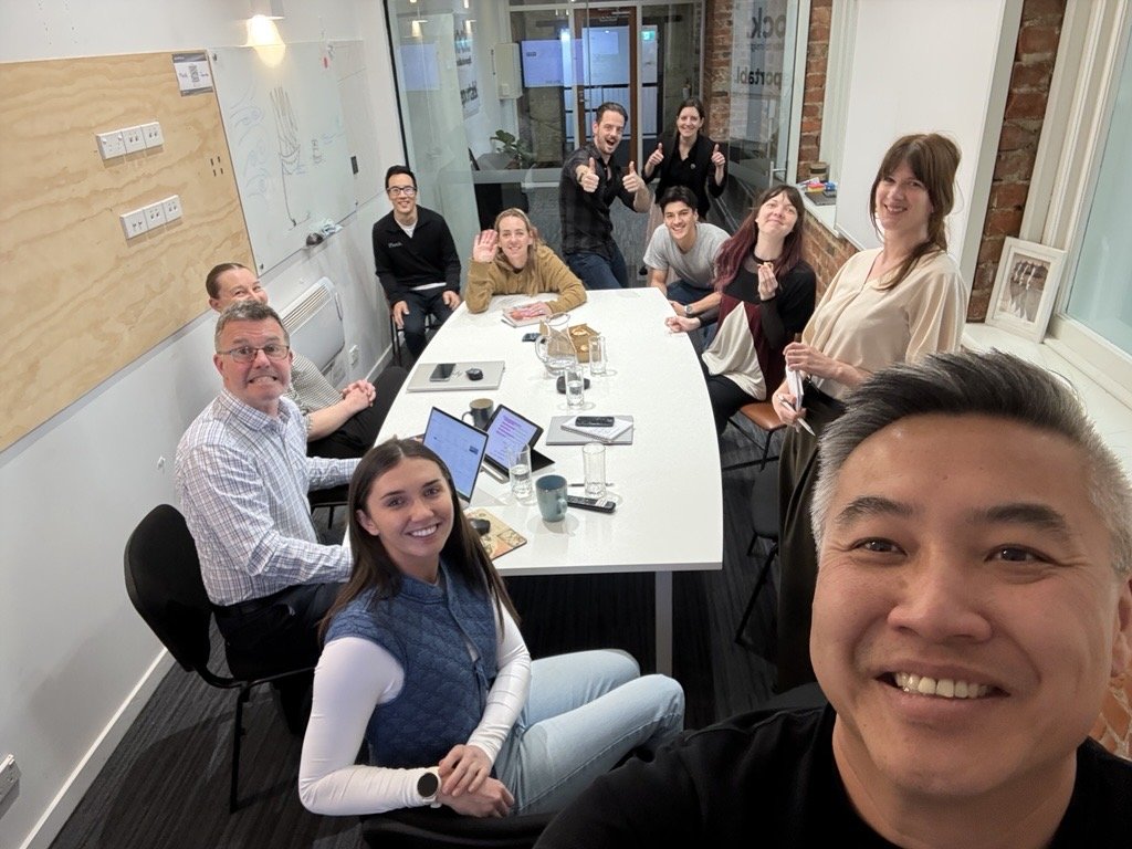 A group of twelve people in a meeting room, smiling and posing for a selfie, with some giving thumbs up and others waving.
