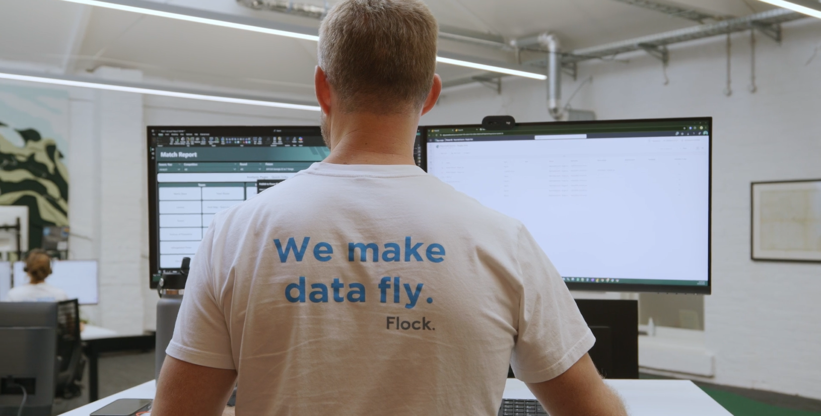 A person with short hair viewed from behind sitting at a desk working on dual computer monitors in an office. The person is wearing a white T-shirt with blue text that reads ‘We make data fly. Flock.’