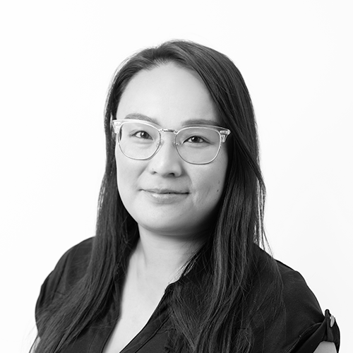 Black and white portrait of a woman with long dark hair wearing glasses and a dark top, smiling slightly against a plain white background.