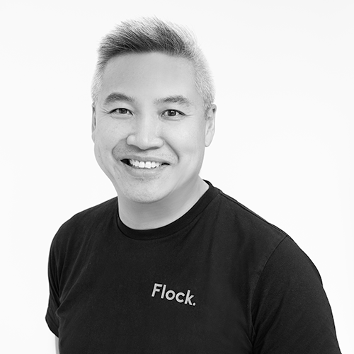 Black and white portrait of a smiling man with short hair wearing a black T-shirt with the word 'Flock.' on it.