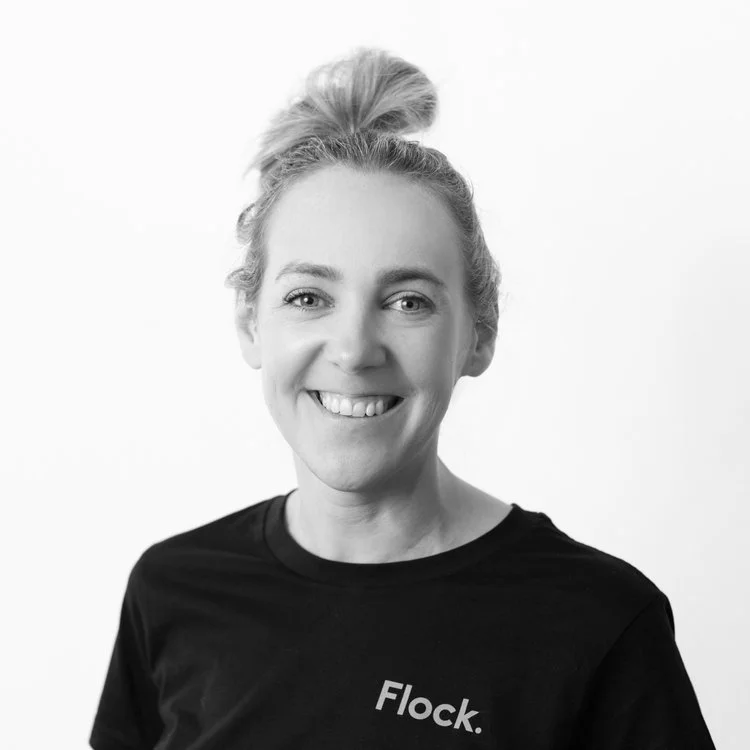 Black and white photo of smiling woman with her hair in a messy bun, wearing a black shirt with the word 'Flock' printed on it.