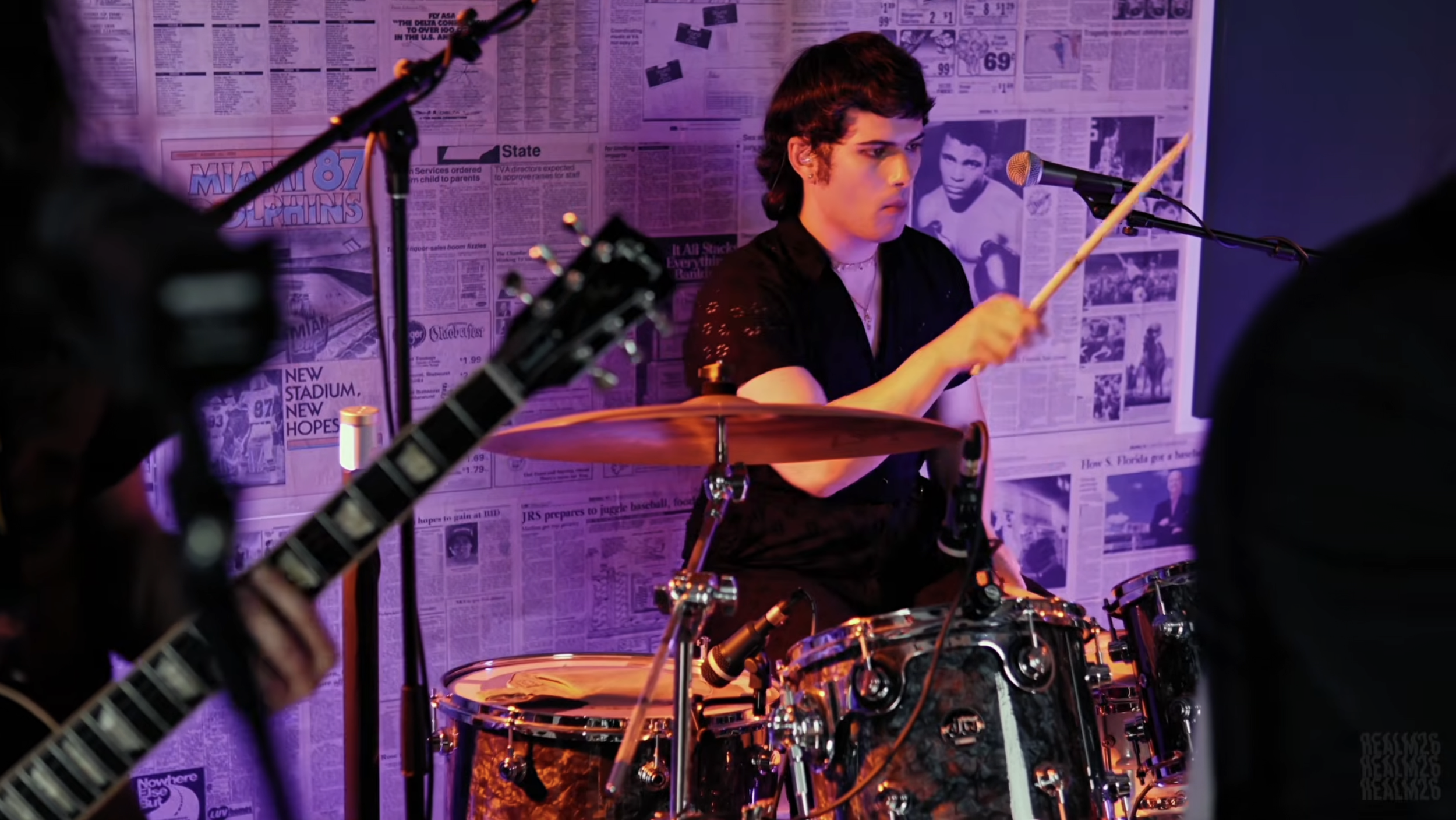 A musician playing drums in a music studio with newspaper-covered walls and purple lighting.